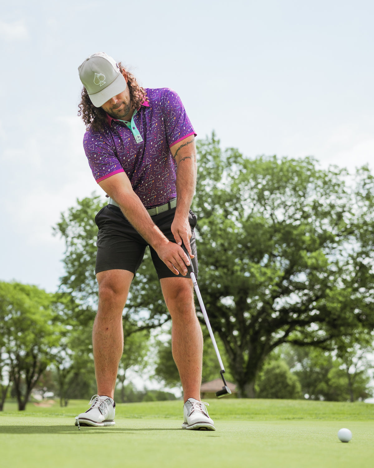 A golfer in a vibrant Eighties Arcade polo and shorts prepares to putt on a sunny green course.