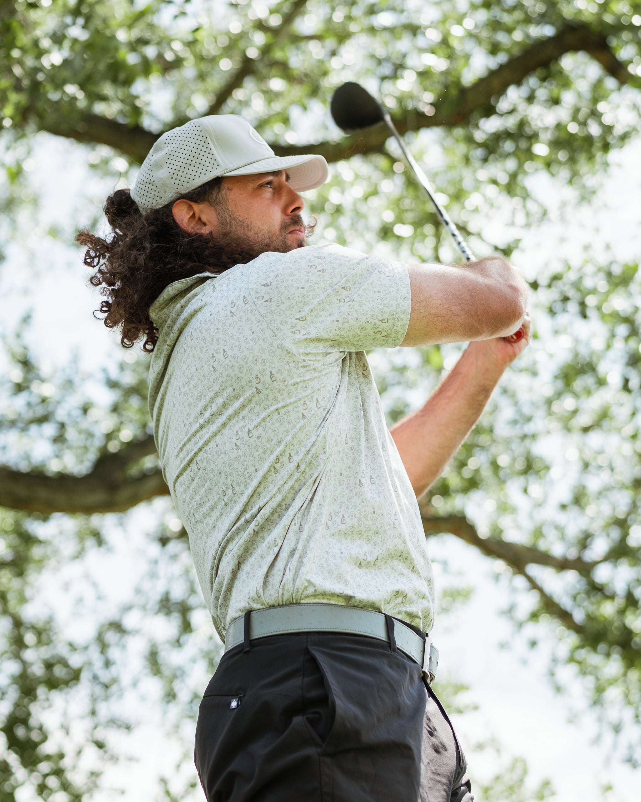 Man swinging a golf club, wearing a stylish patterned shirt and cap, surrounded by trees on a sunny day.