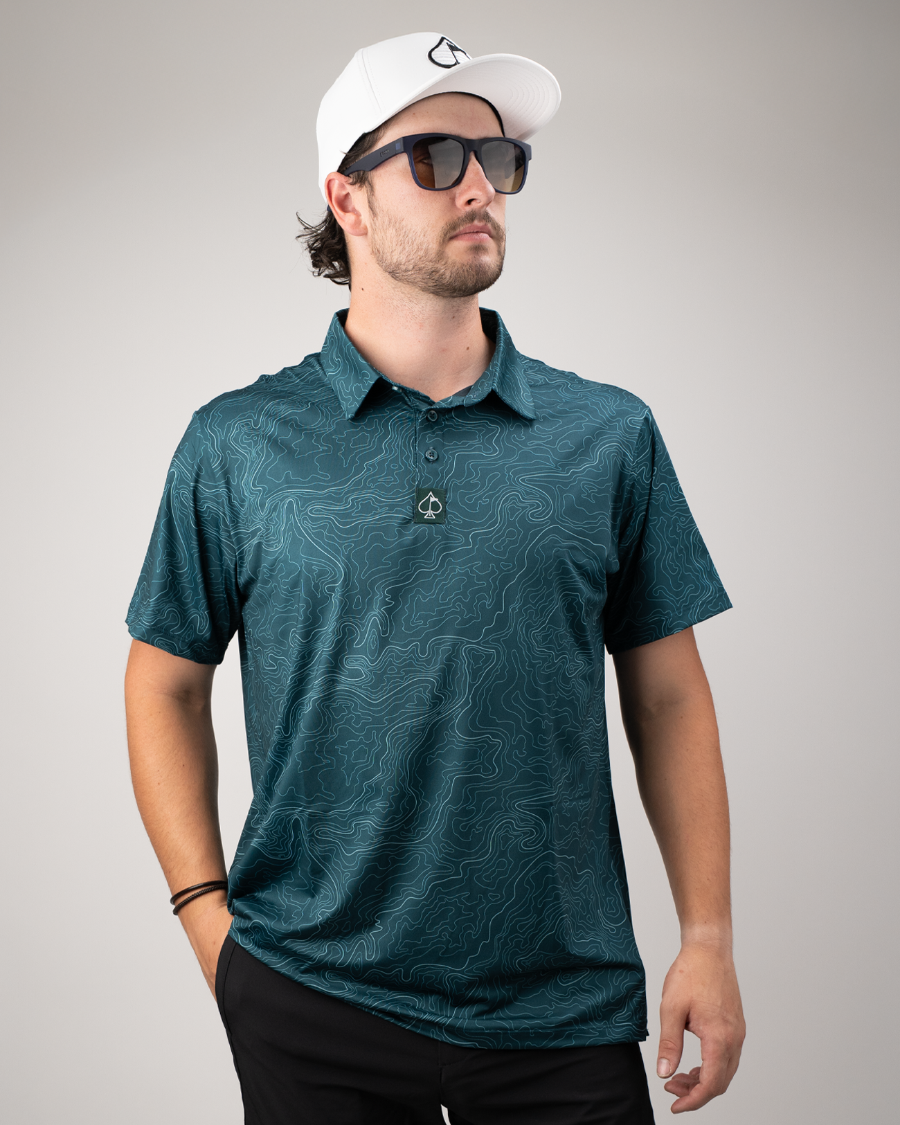 Men's Terrain golf polo in teal with a topographic design, worn by a model with sunglasses and a white cap.