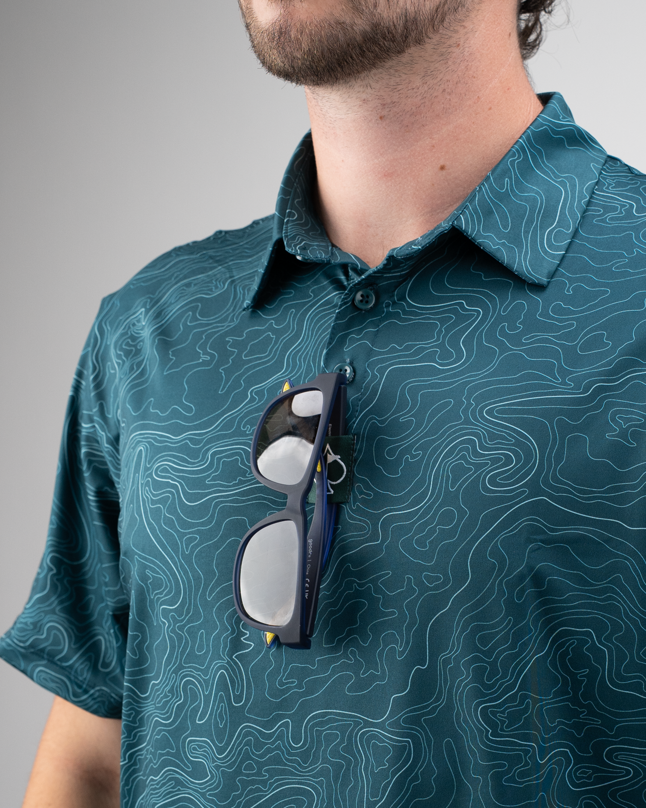 Close-up of a man wearing a teal golf polo with a topographic pattern, sunglasses resting on the collar.