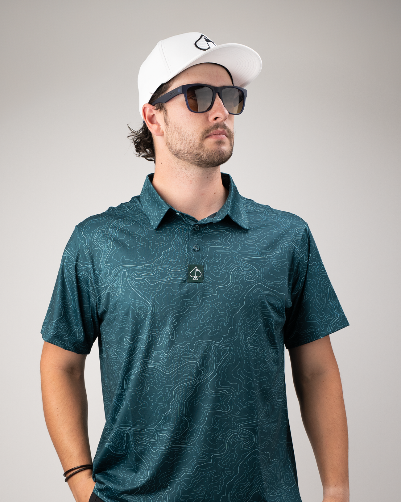 Man wearing a teal golf polo with a terrain pattern, sunglasses, and a white cap, posing against a neutral background.