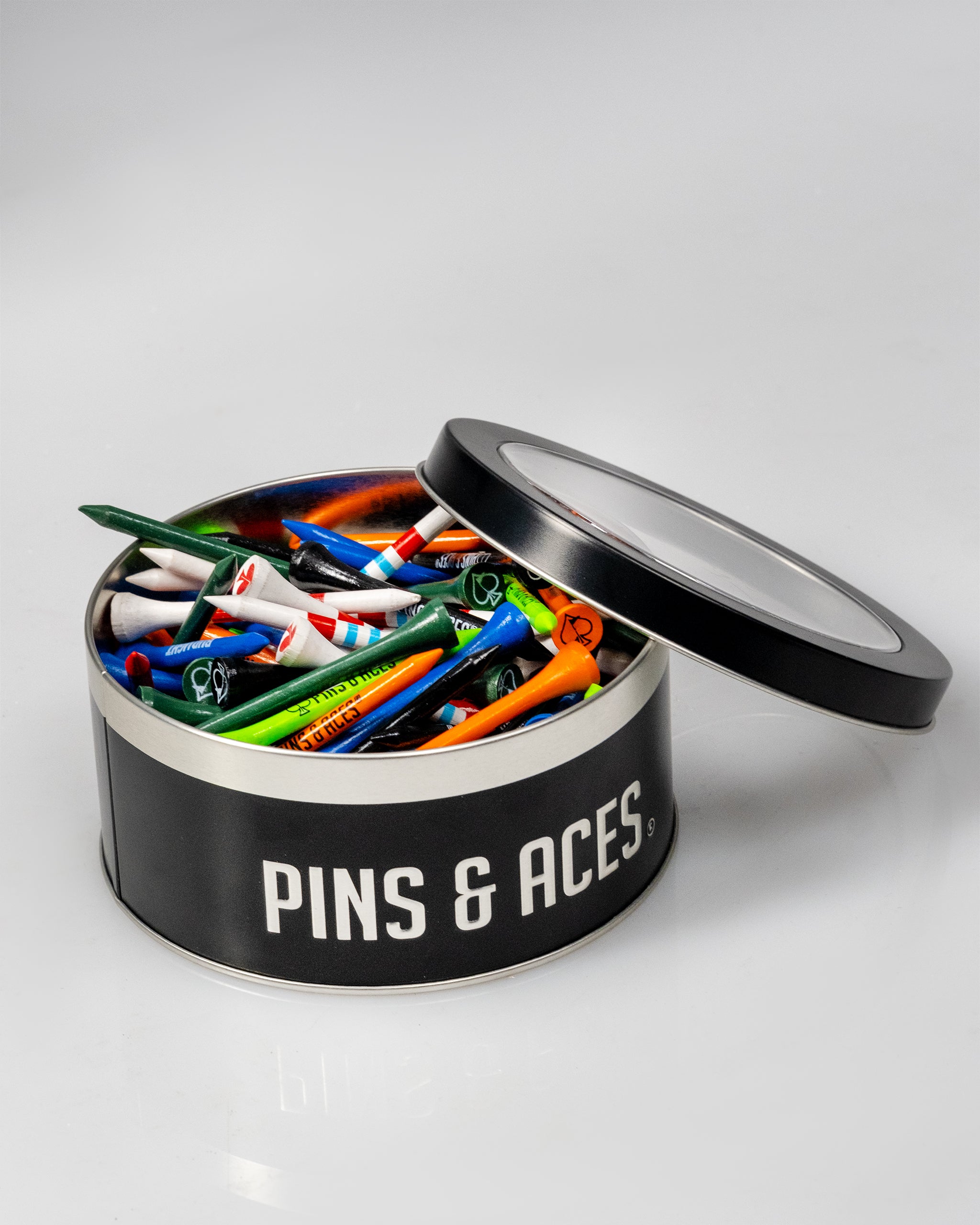 Round black tin labeled "Pins & Aces" filled with colorful golf tees, some with brand logos, on a plain white background.