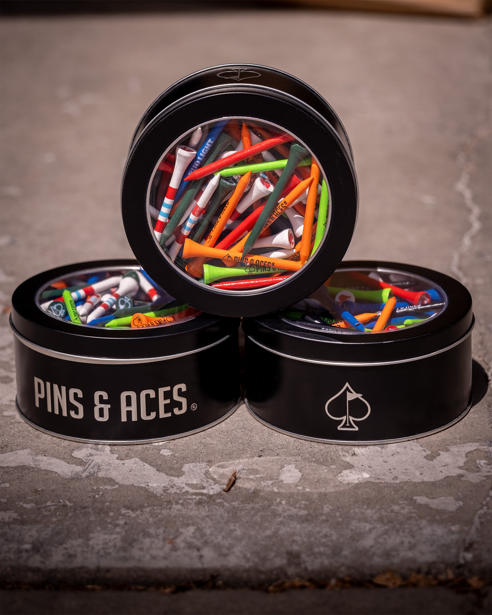 Three black tins labeled "Pins & Aces" filled with colorful golf tees, stacked on a concrete surface.