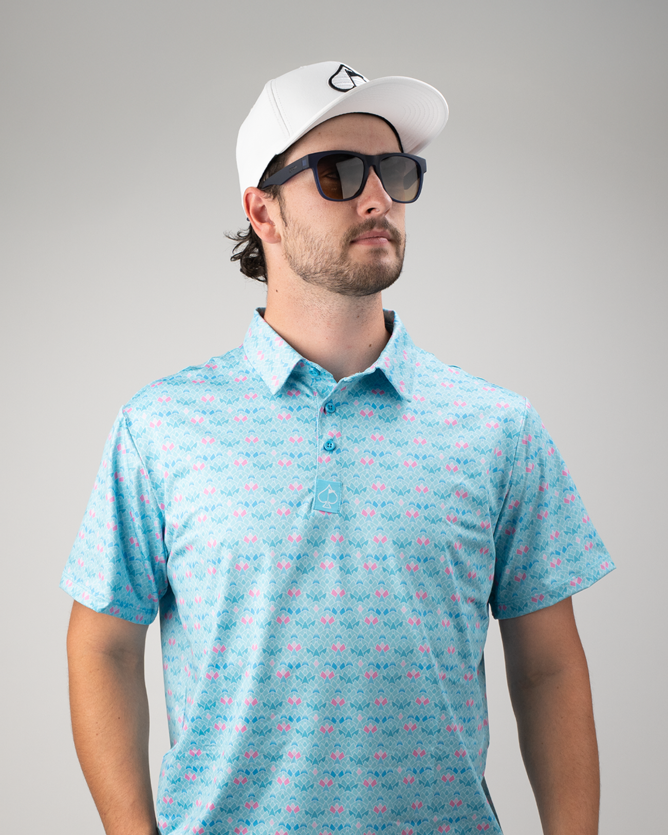 Man wearing a light blue golf polo with a pink triangle bloom pattern, sunglasses, and a white cap, posing confidently.