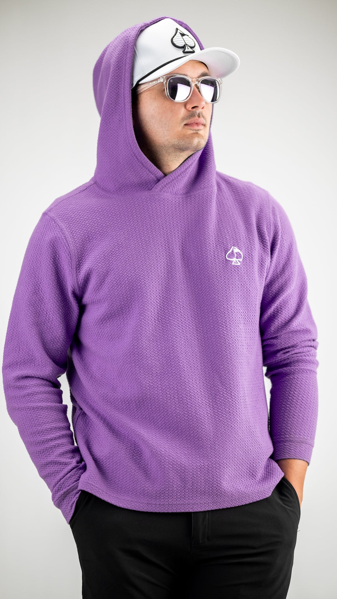 Male golfer in a purple textured hoodie with a spade logo, wearing a white cap with spade symbol, reflective sunglasses, a...