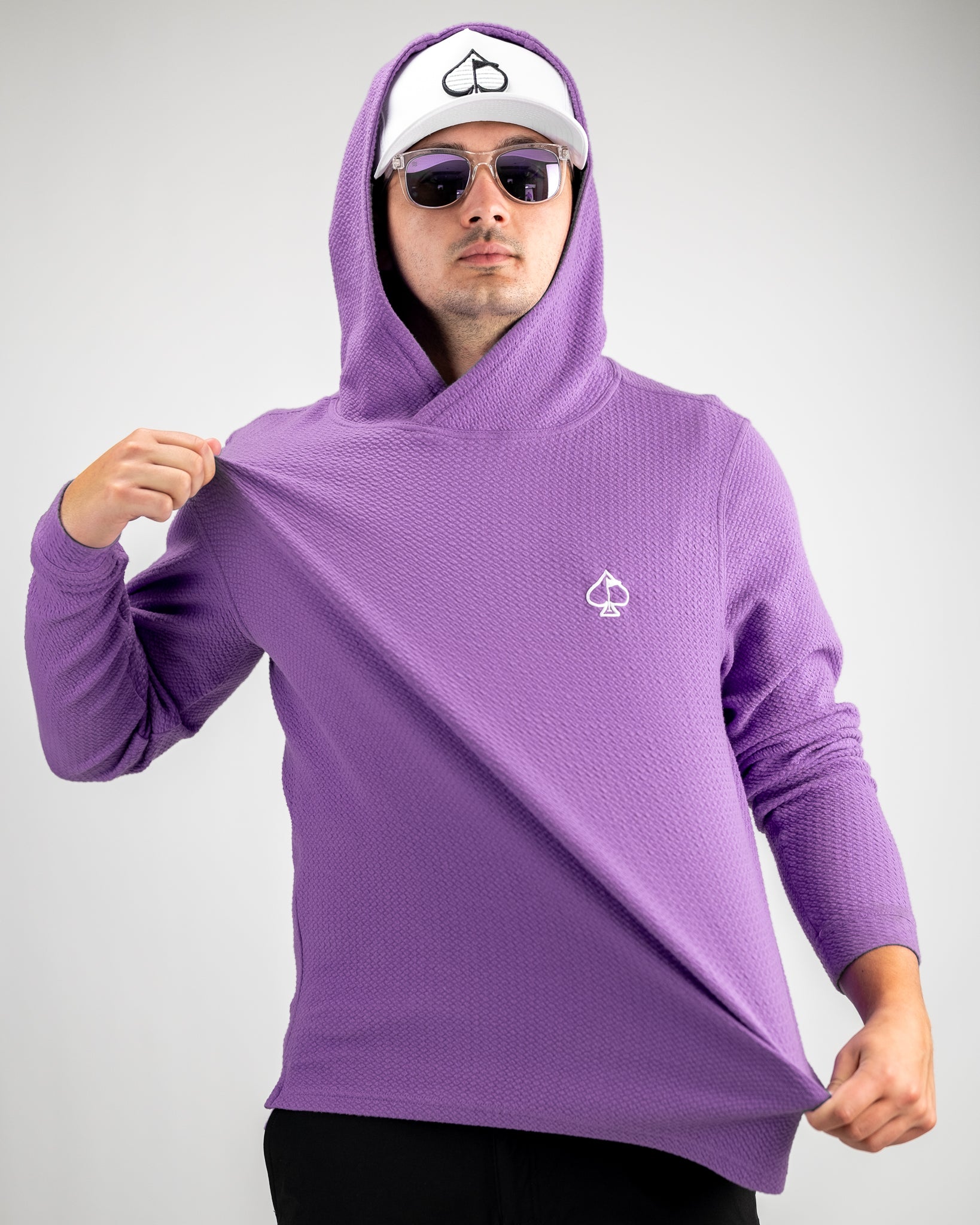 Man wearing a purple textured hoodie with a white cap featuring a black spade logo, and sunglasses, showcasing bold golf a...
