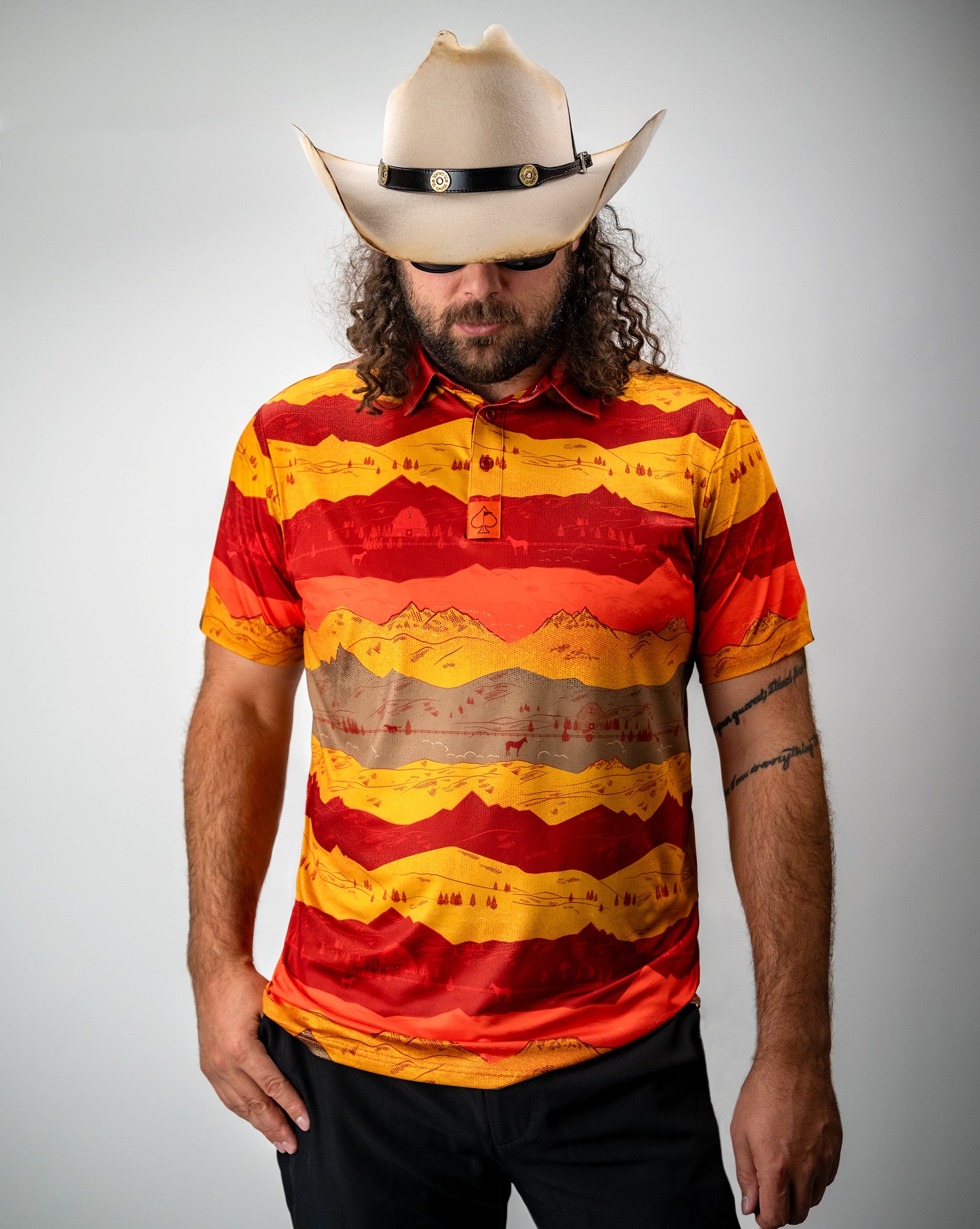 Man wearing a colorful, bold mountain landscape golf shirt in red, yellow, and orange, with a cowboy hat and sunglasses.