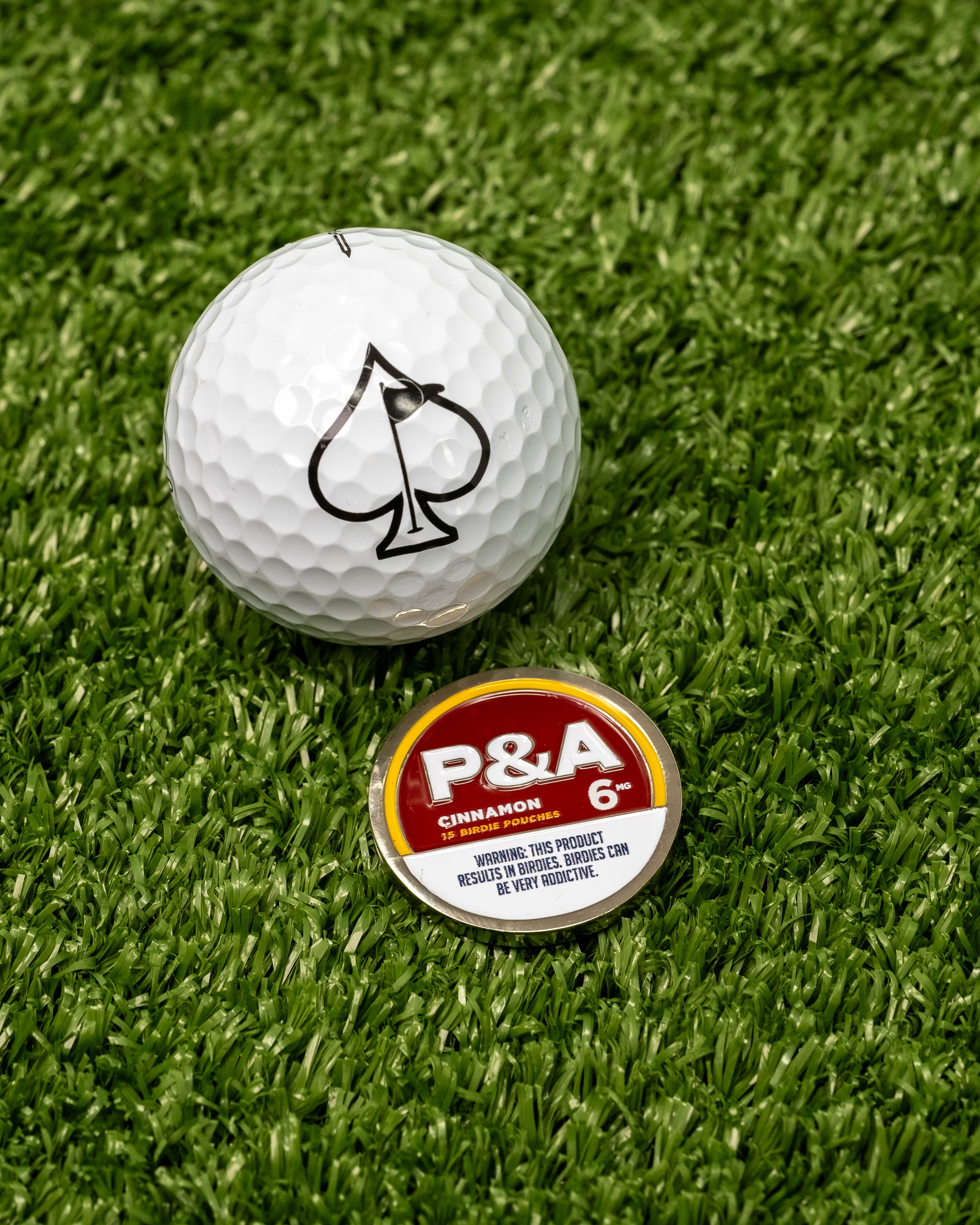 "6 Milly Cinnamon Ball Marker next to a golf ball with an ace of spades logo on green turf.