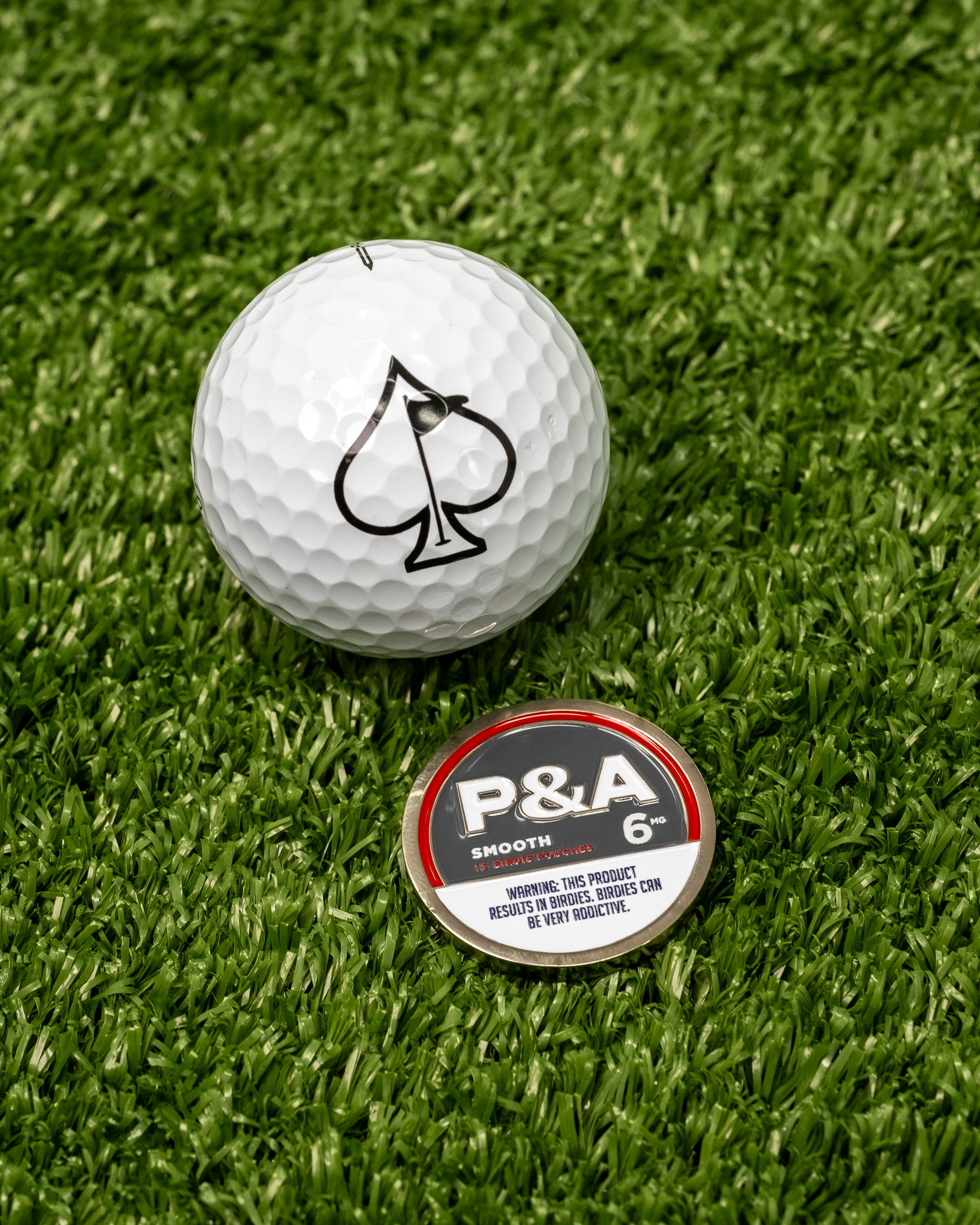 White golf ball with black spade logo next to a round marker labeled "P&A 6" on green turf.