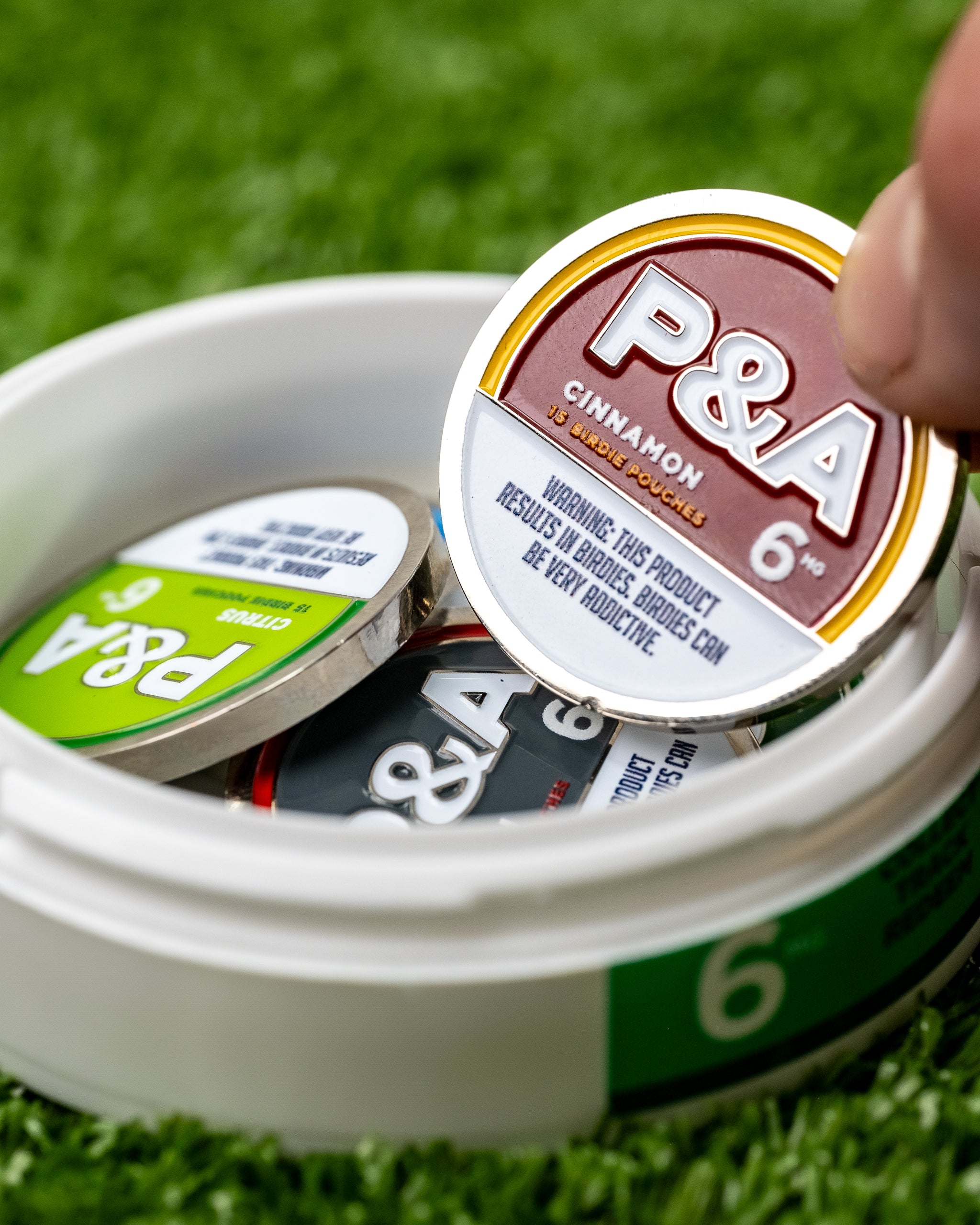 "Close-up of 6 Milly Cinnamon Ball Marker being lifted from a container on green turf.