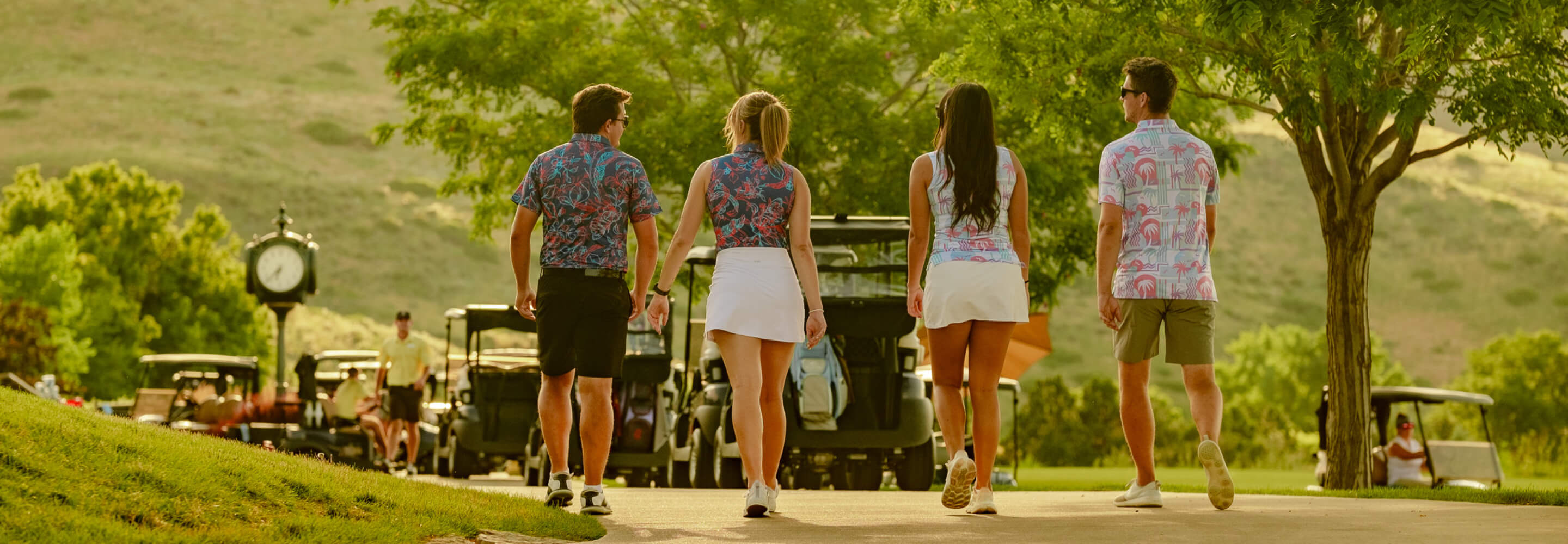 Group of golfers in colorful, tropical-themed shirts walking on a golf course with carts in the background.