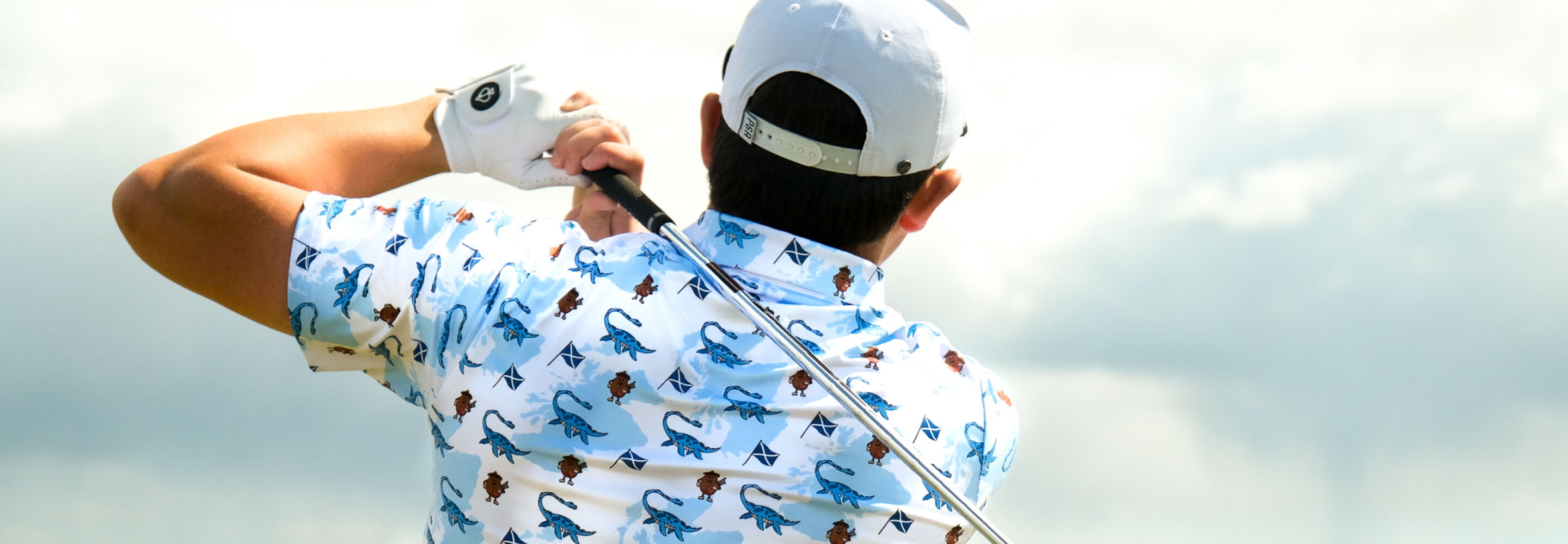 Male golfer in a blue patterned shirt and white cap swings on a cloudy day, showcasing bold golf apparel and accessories.