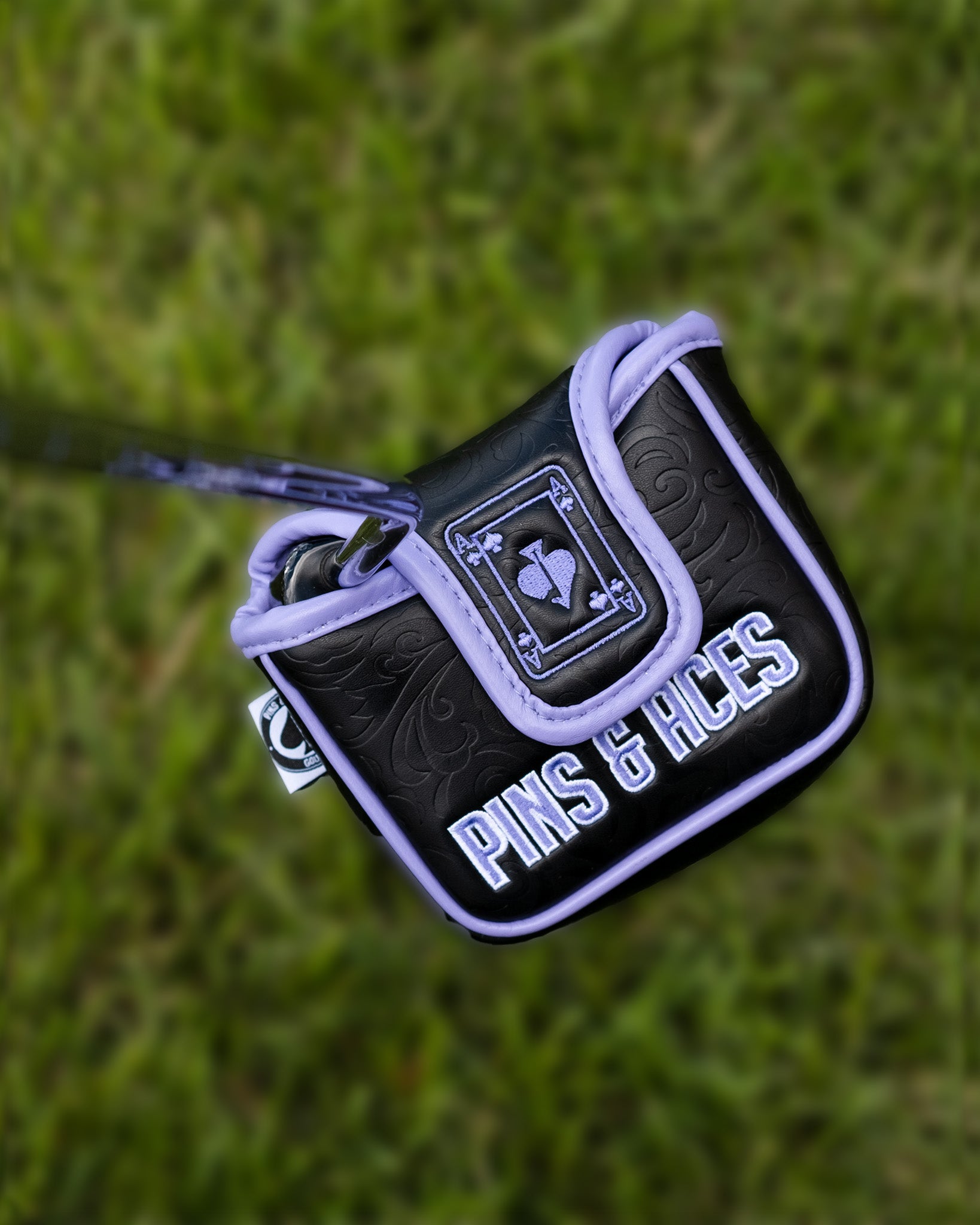 Alt text: "Ace of Spades mallet putter cover by Pins & Aces, featuring a detailed embroidered design on black and purple padded fabric, set against a grassy background."