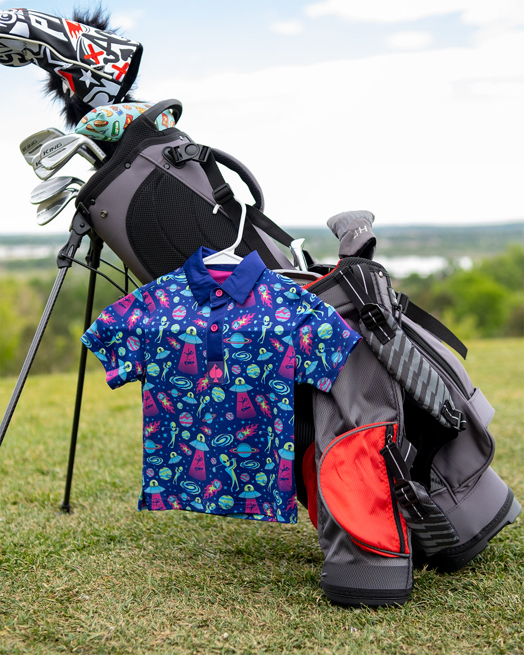 Youth Alien Invasion Polo featuring a playful alien graphic, displayed hanging on a bag against a neutral background.