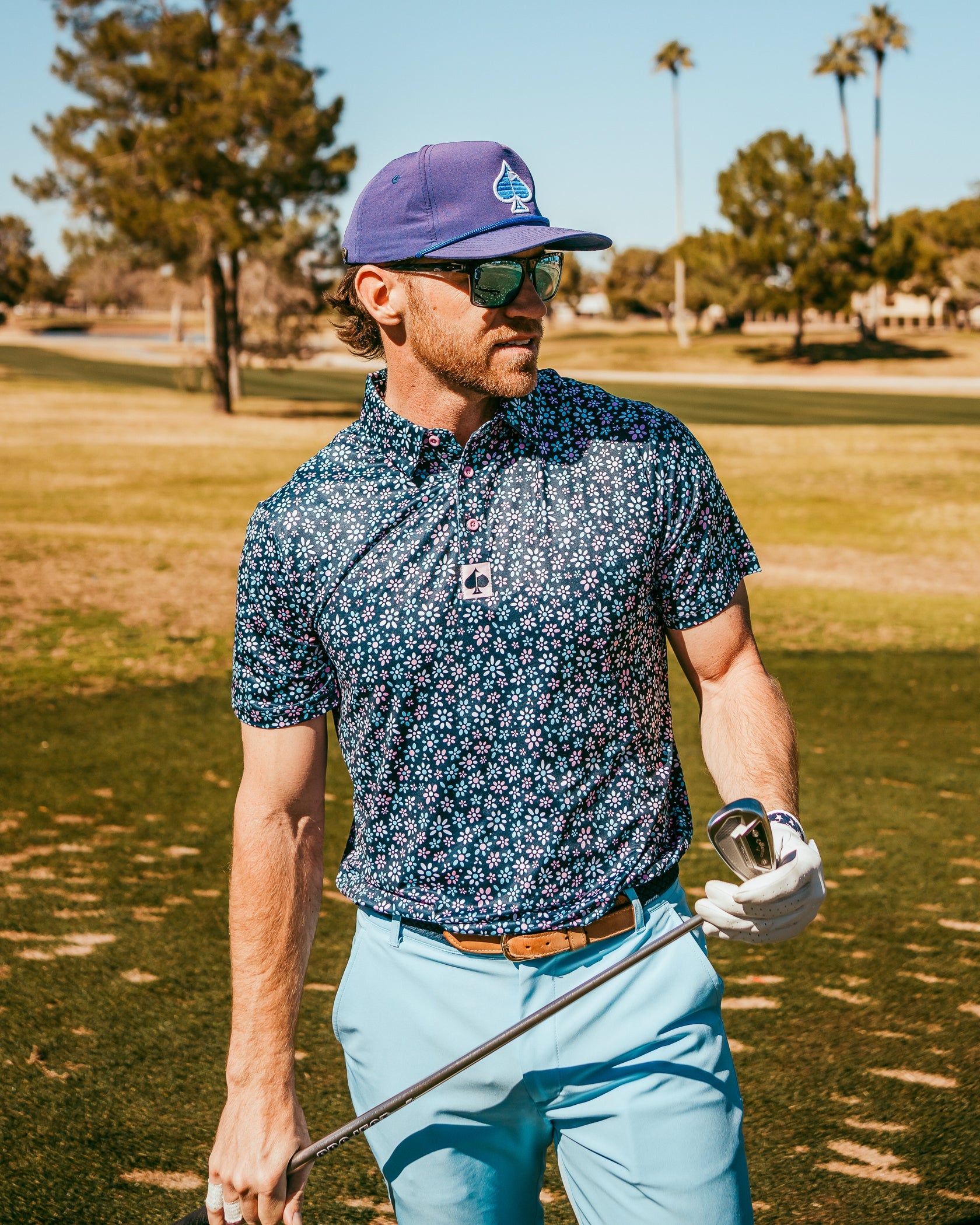 A golfer wearing a floral-patterned polo shirt and light blue pants stands on the course, holding a club.