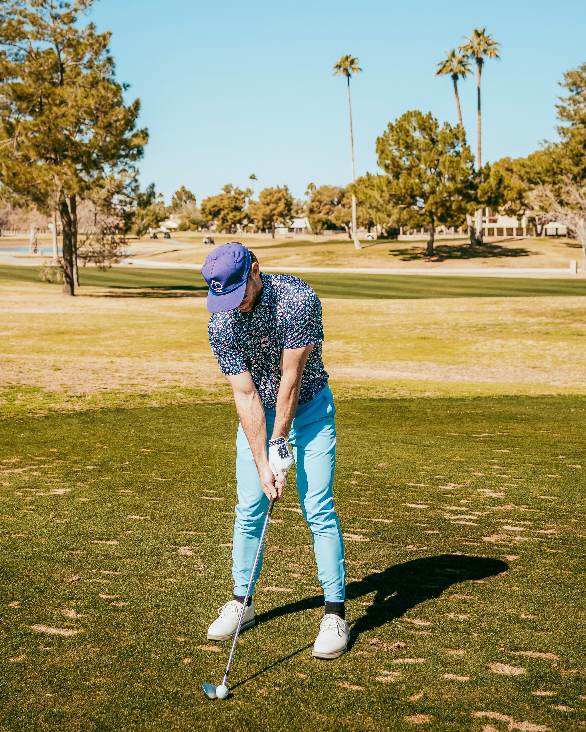 A golfer in a floral shirt and light blue pants prepares to swing on a sunny golf course surrounded by palm trees.