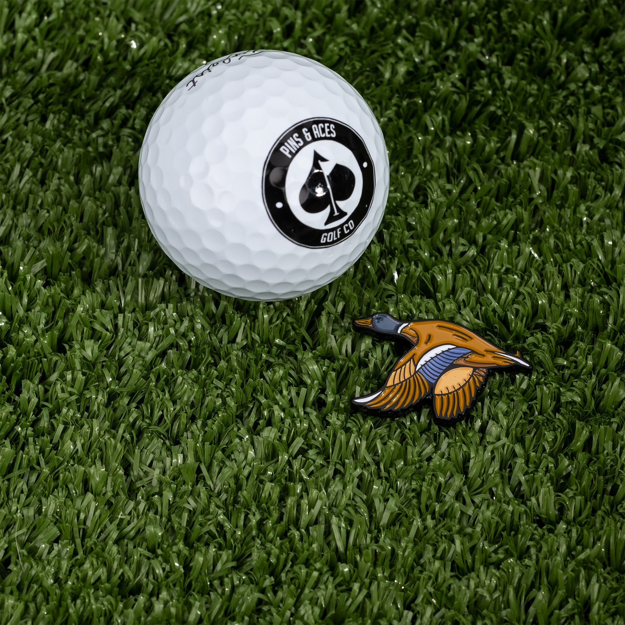 Flying Mallard ball marker beside a golf ball on green turf, showcasing a vibrant duck design for stylish marking.