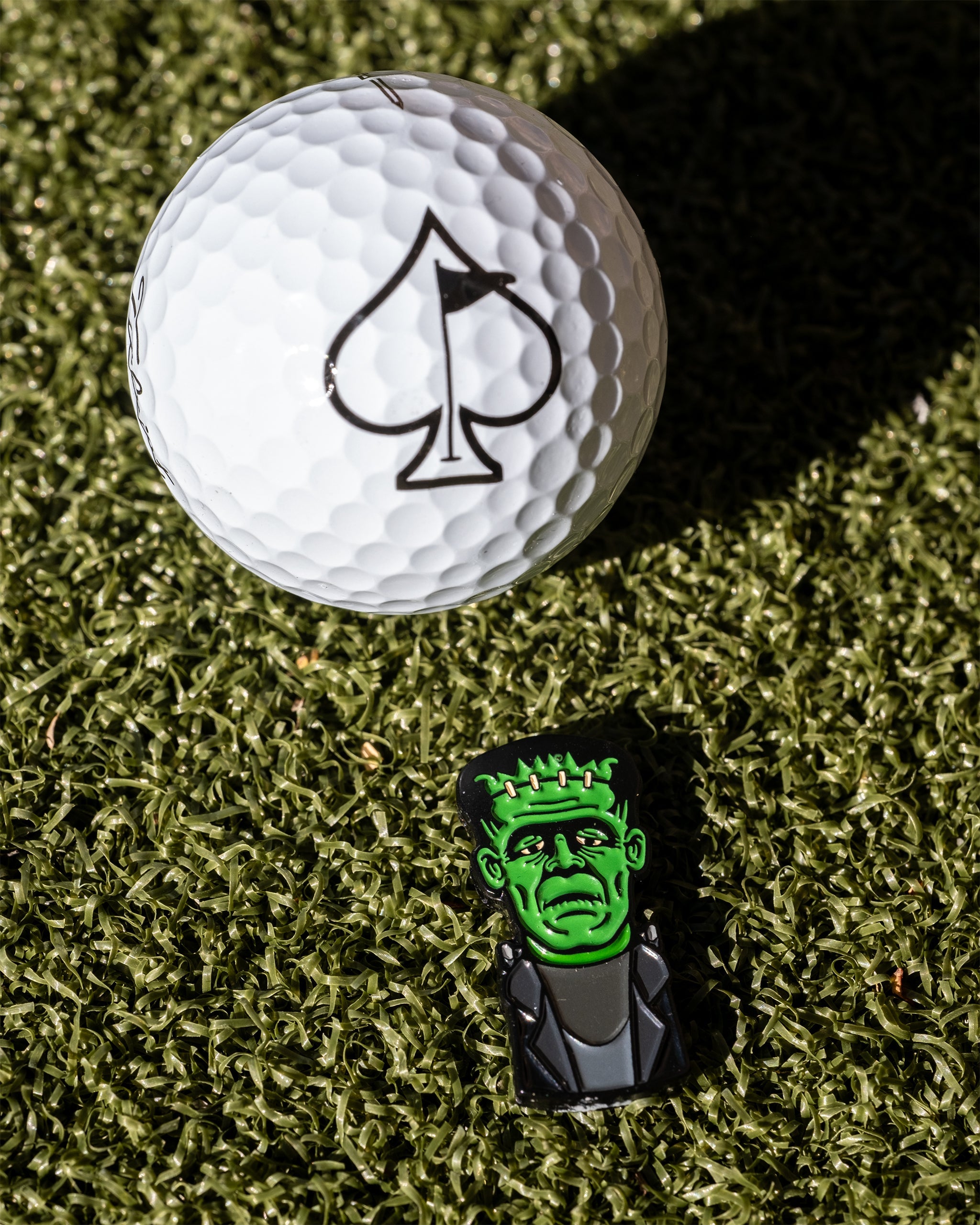 Frankenstein-themed golf ball marker beside a white golf ball with an ace of spades logo on a green turf background.