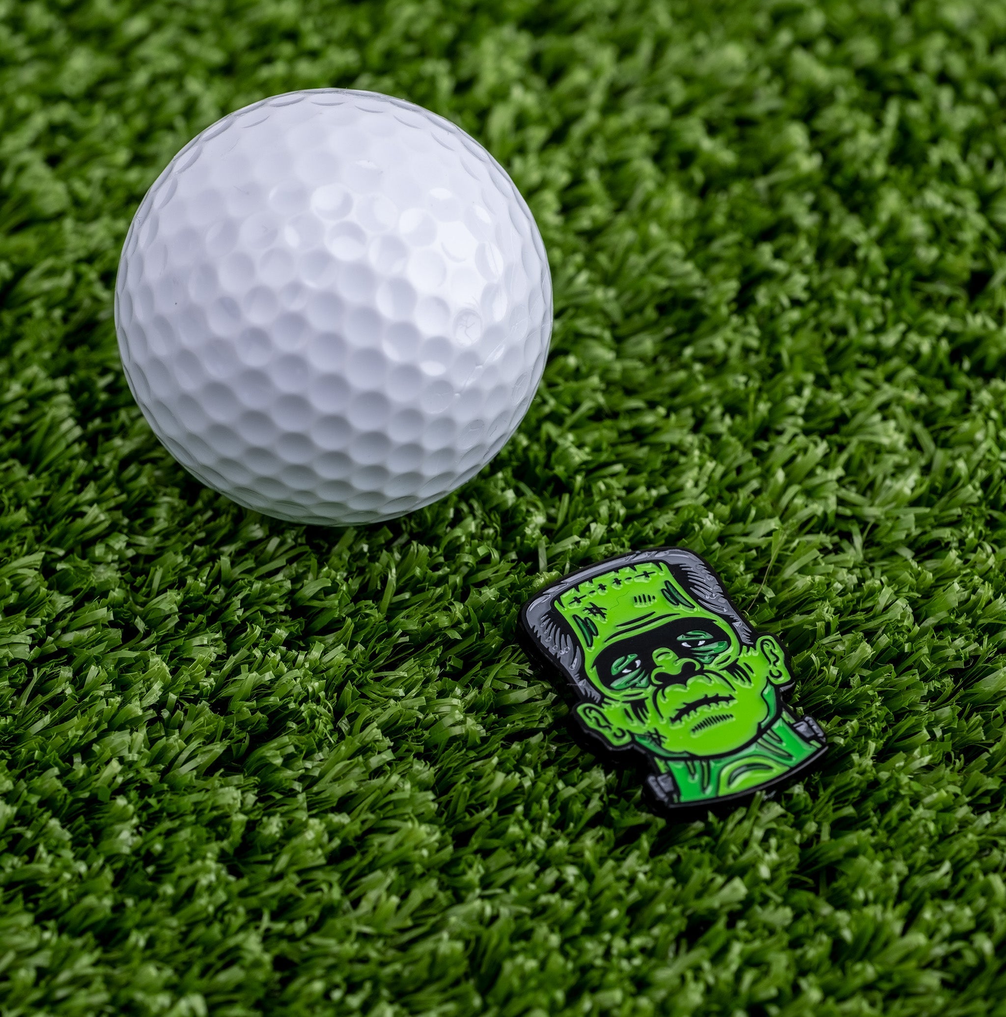 Frankenstein-themed golf ball marker next to a white golf ball on green turf, perfect for adding personality to your game.
