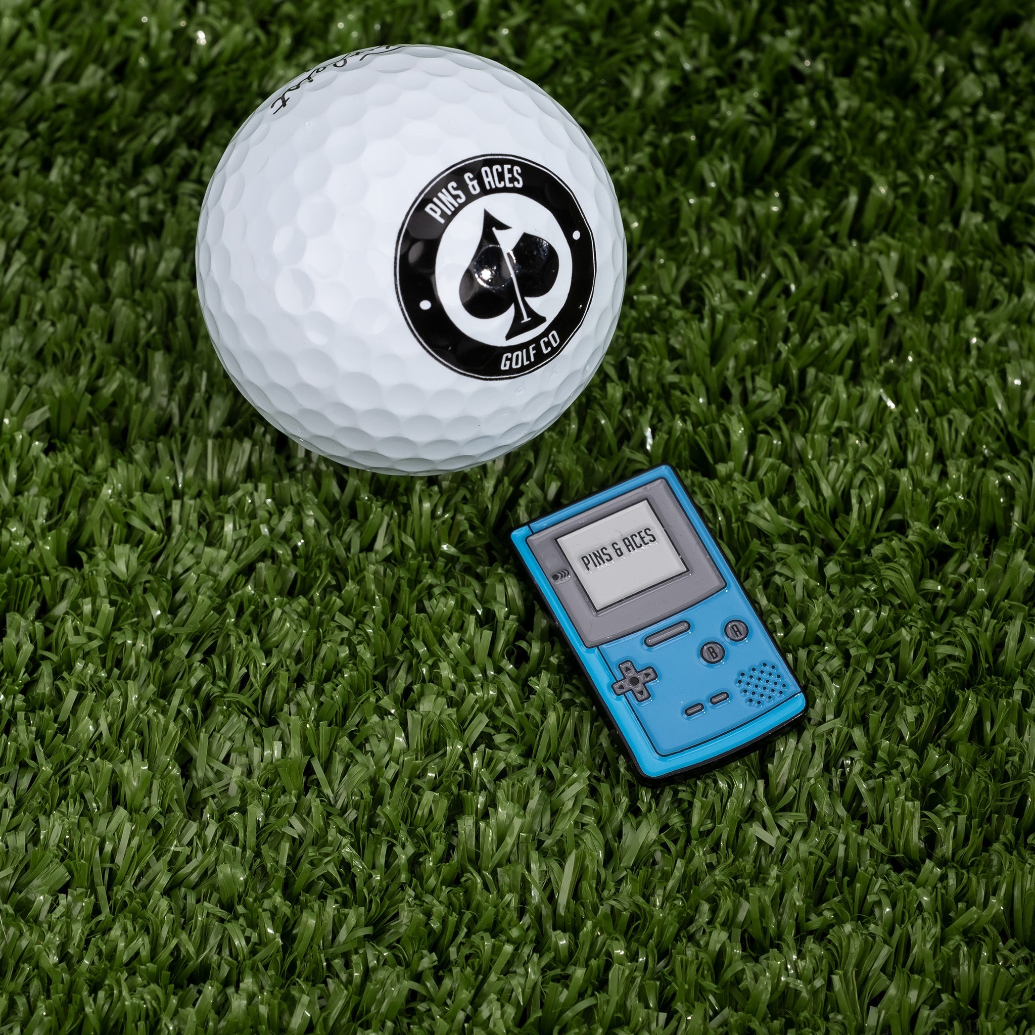 Game Boy-inspired ball marker in blue next to a white golf ball with a logo on a grassy surface.