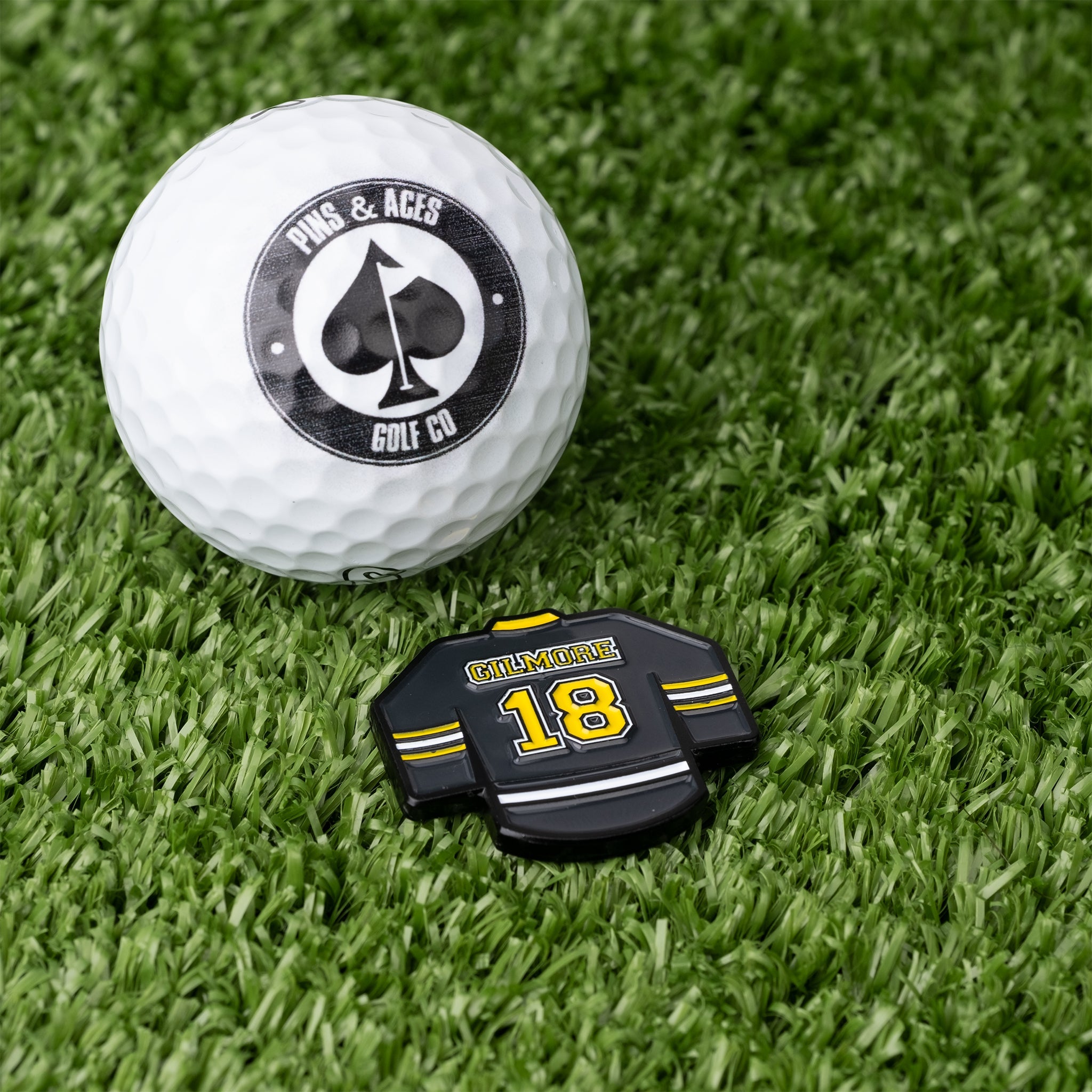 Gilmore jersey ball marker in black and yellow next to a white golf ball on green turf, perfect for stylish golfers.