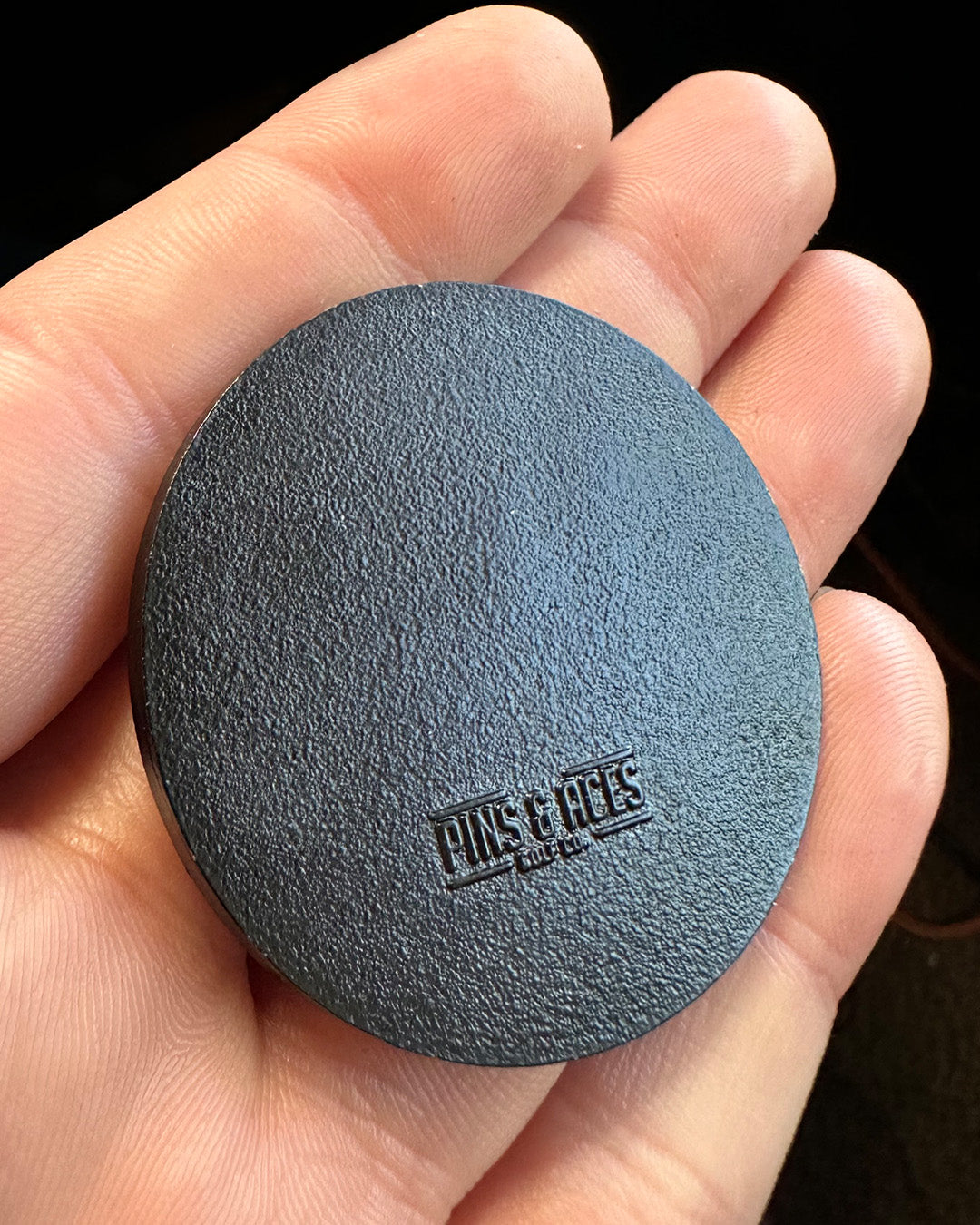 Black golf ball marker held in a hand, featuring a textured surface and the Pins and Aces logo. Ideal for golfers.