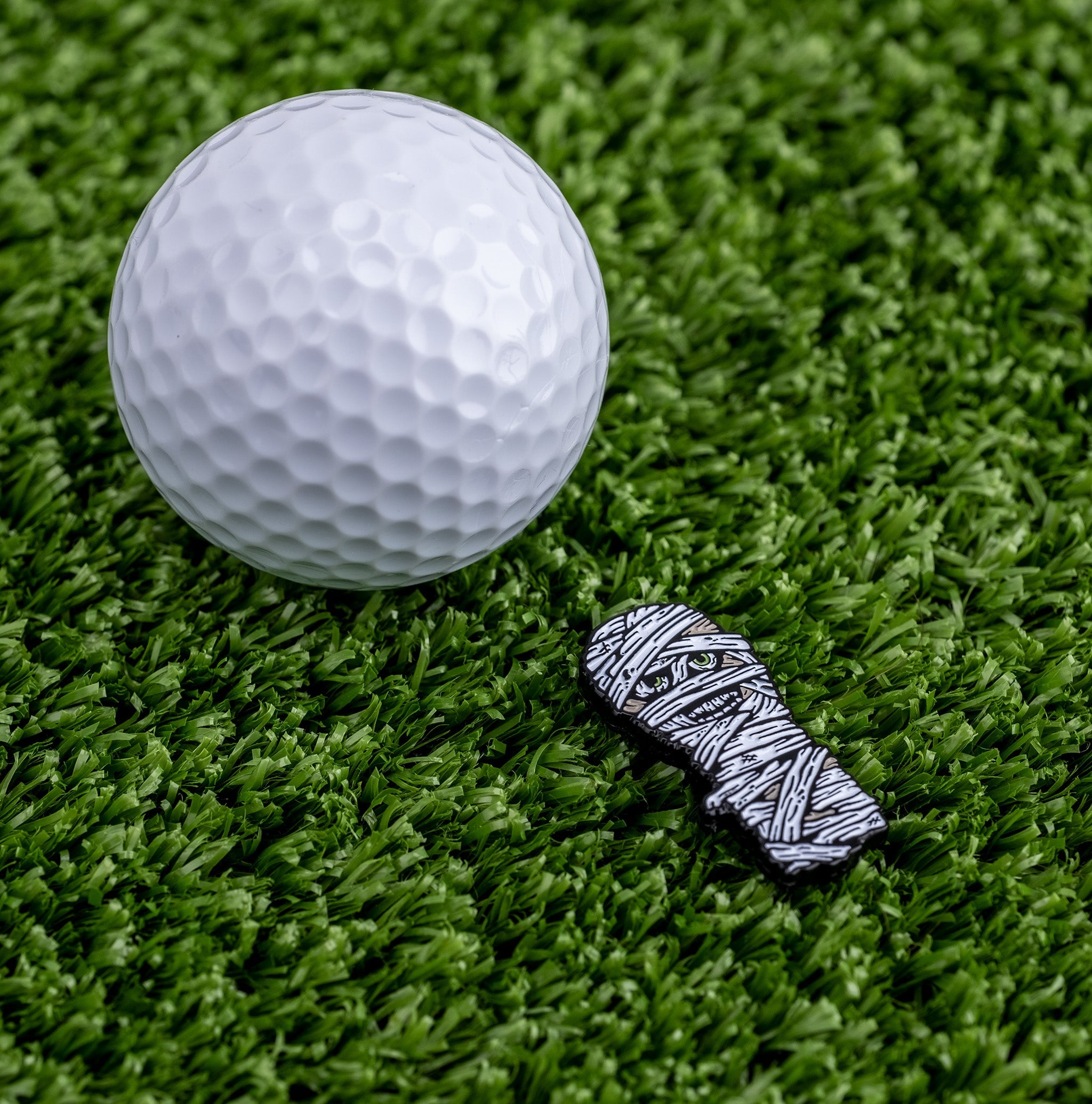 Mummy ball marker beside a white golf ball on green turf, featuring a fun, spooky design for golfers.