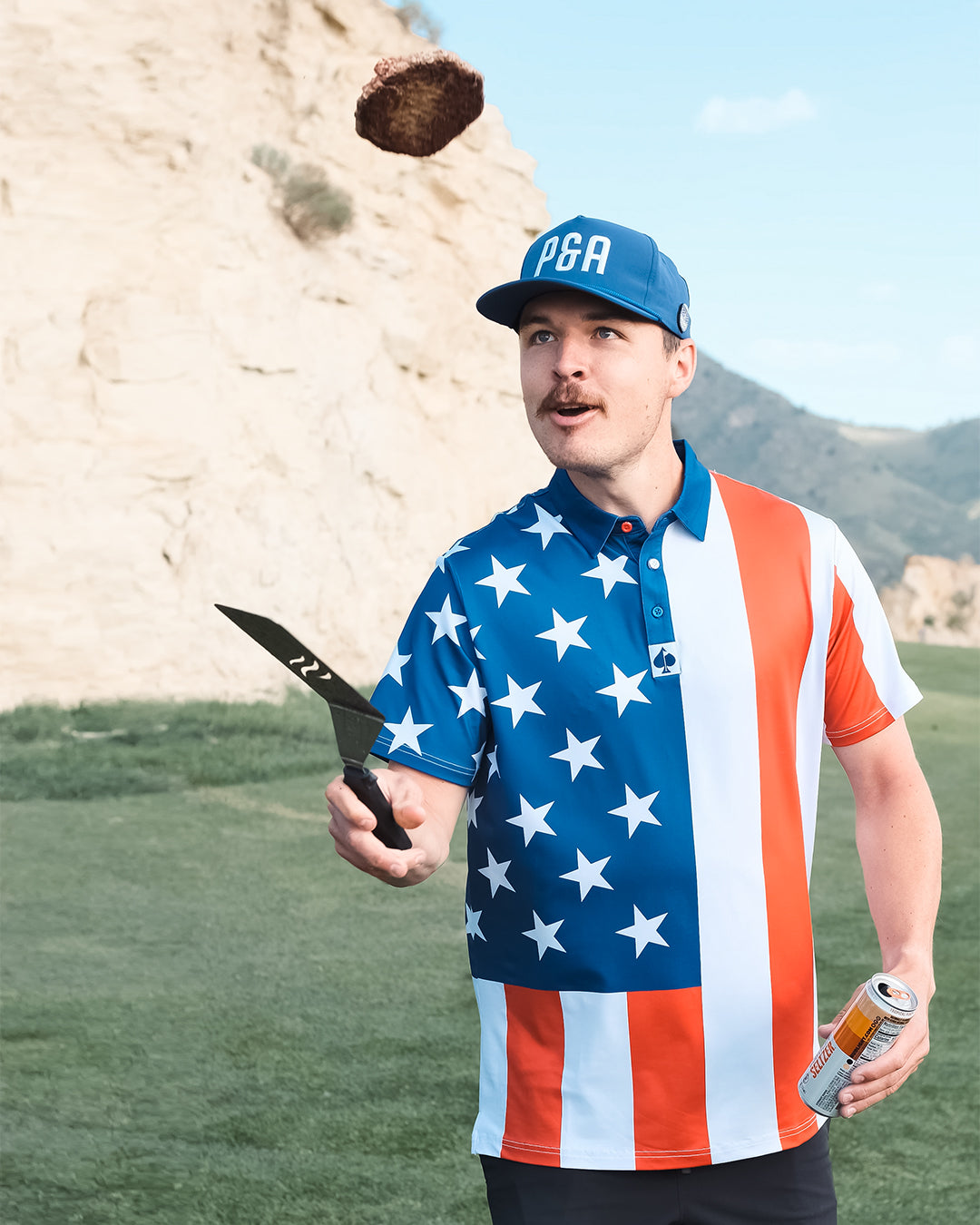 A golfer wearing a patriotic red, white, and blue polo shirt with stars tosses a golf club while standing on a course.