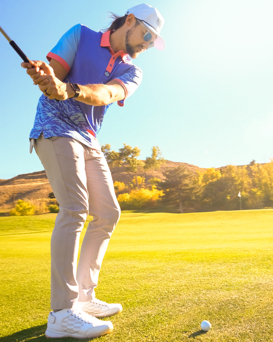 Male golfer in colorful polo and beige pants, preparing to hit on a sunny golf course with scenic hills in the background.