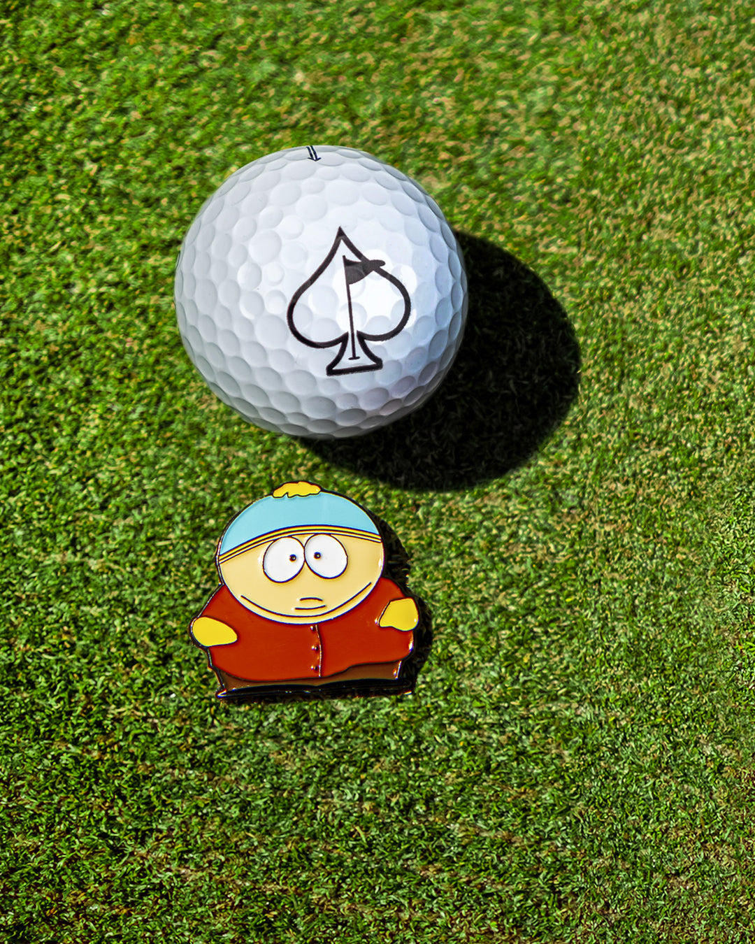 South Park Cartman ball marker beside a golf ball on green grass, perfect for adding personality to your golf game.