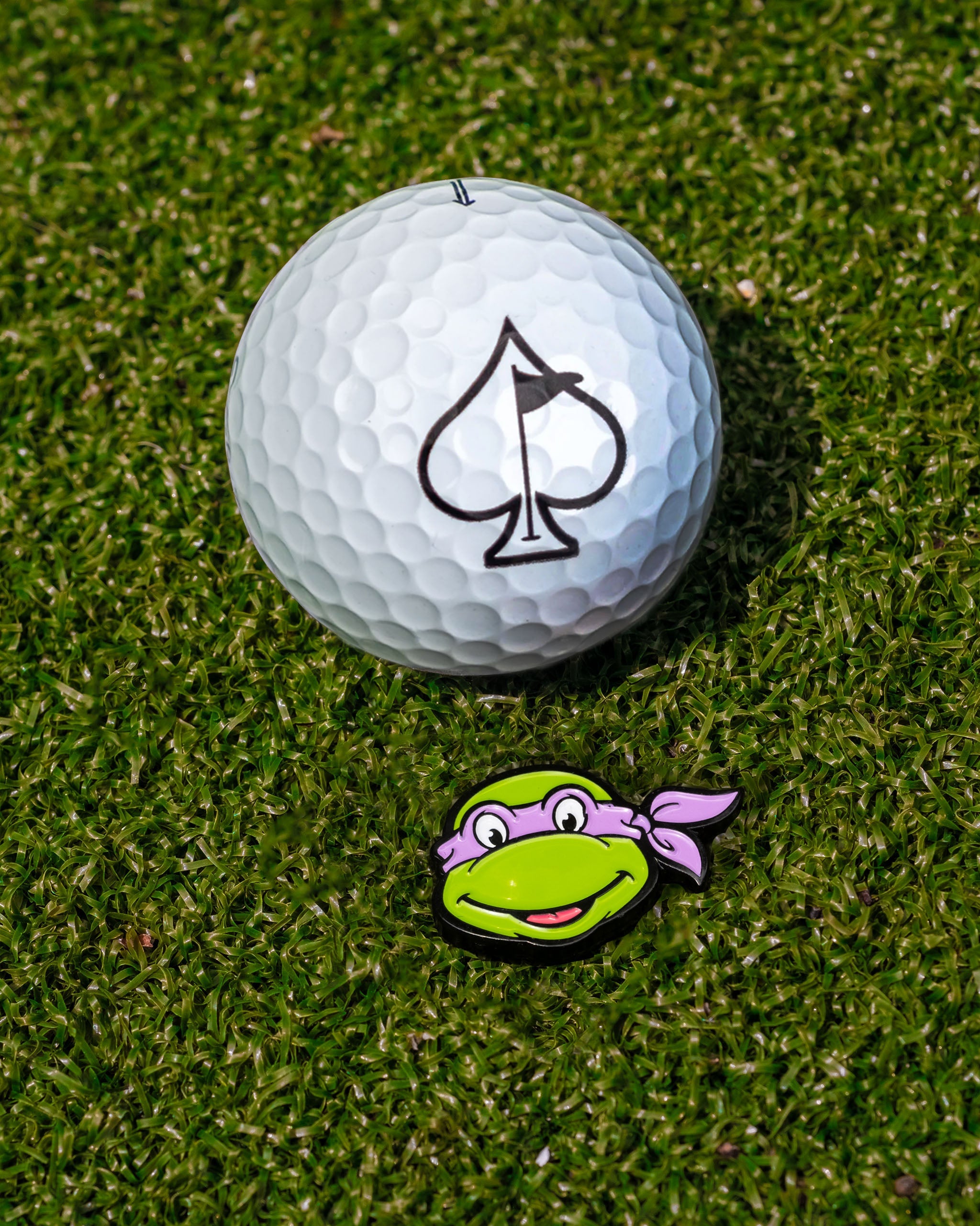 TMNT Donatello ball marker beside a golf ball with a spade logo, set on green grass. Perfect for unique golf accessories.