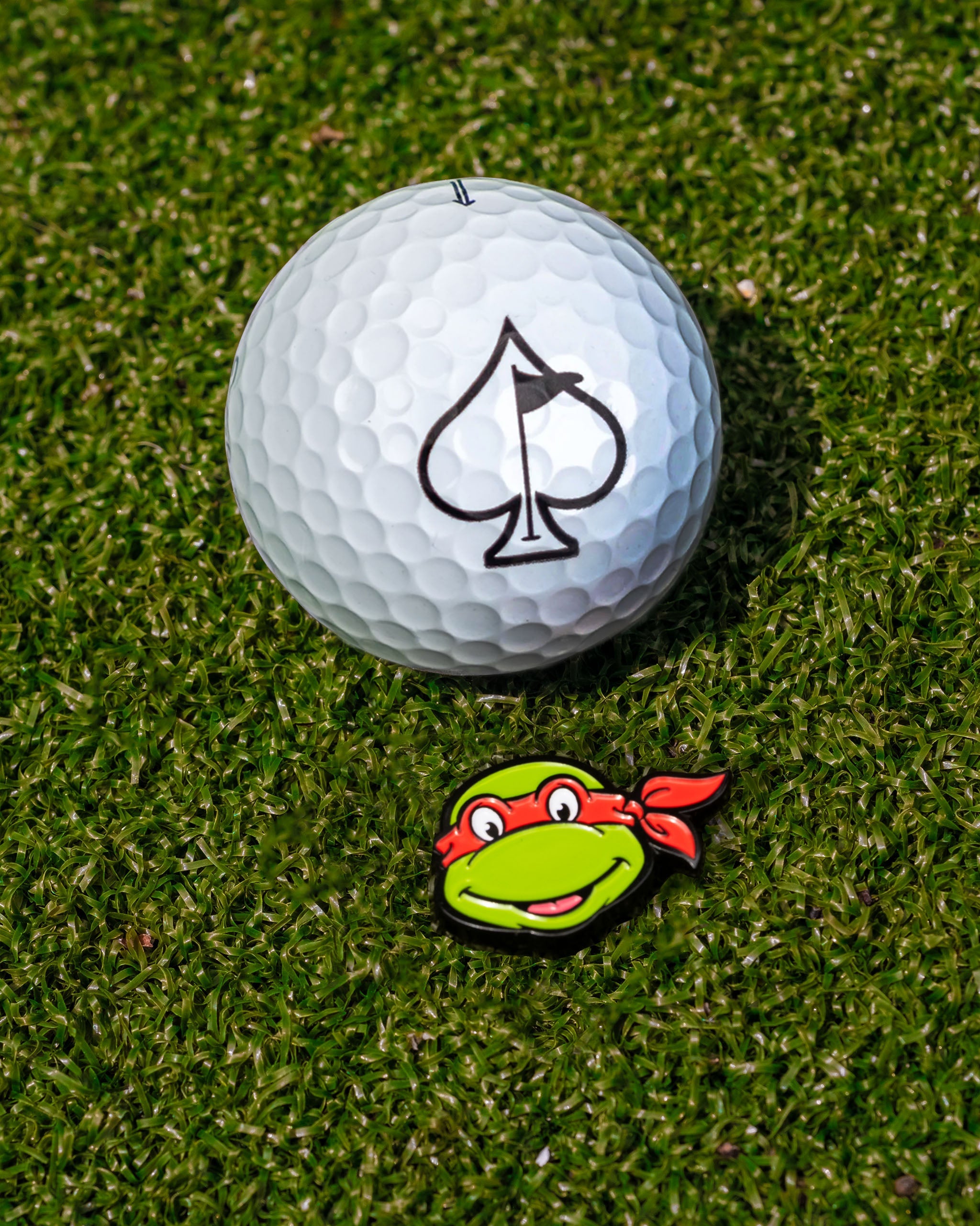 TMNT Raphael ball marker next to a golf ball with an ace of spades logo on a green turf background.