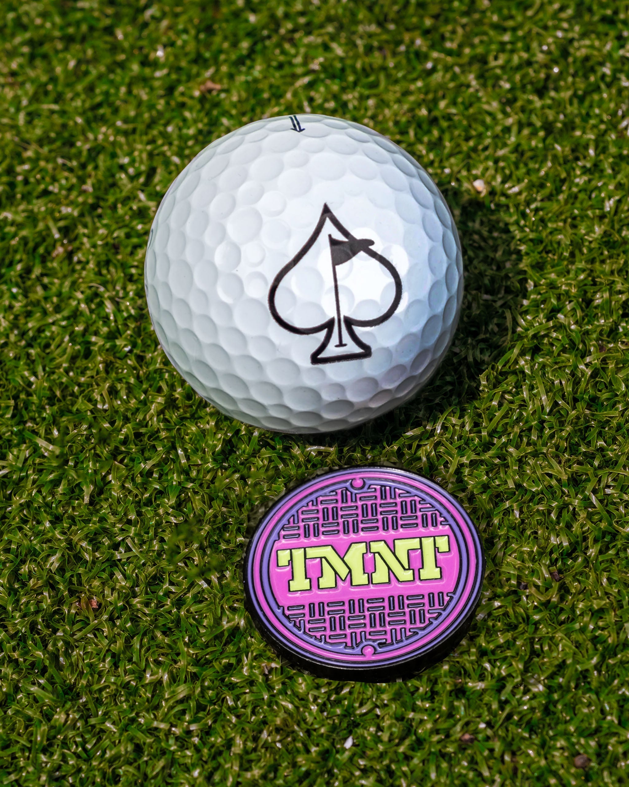 TMNT sewer cover ball marker beside a golf ball with a spade logo, set on green grass. Perfect for unique golf accessories.