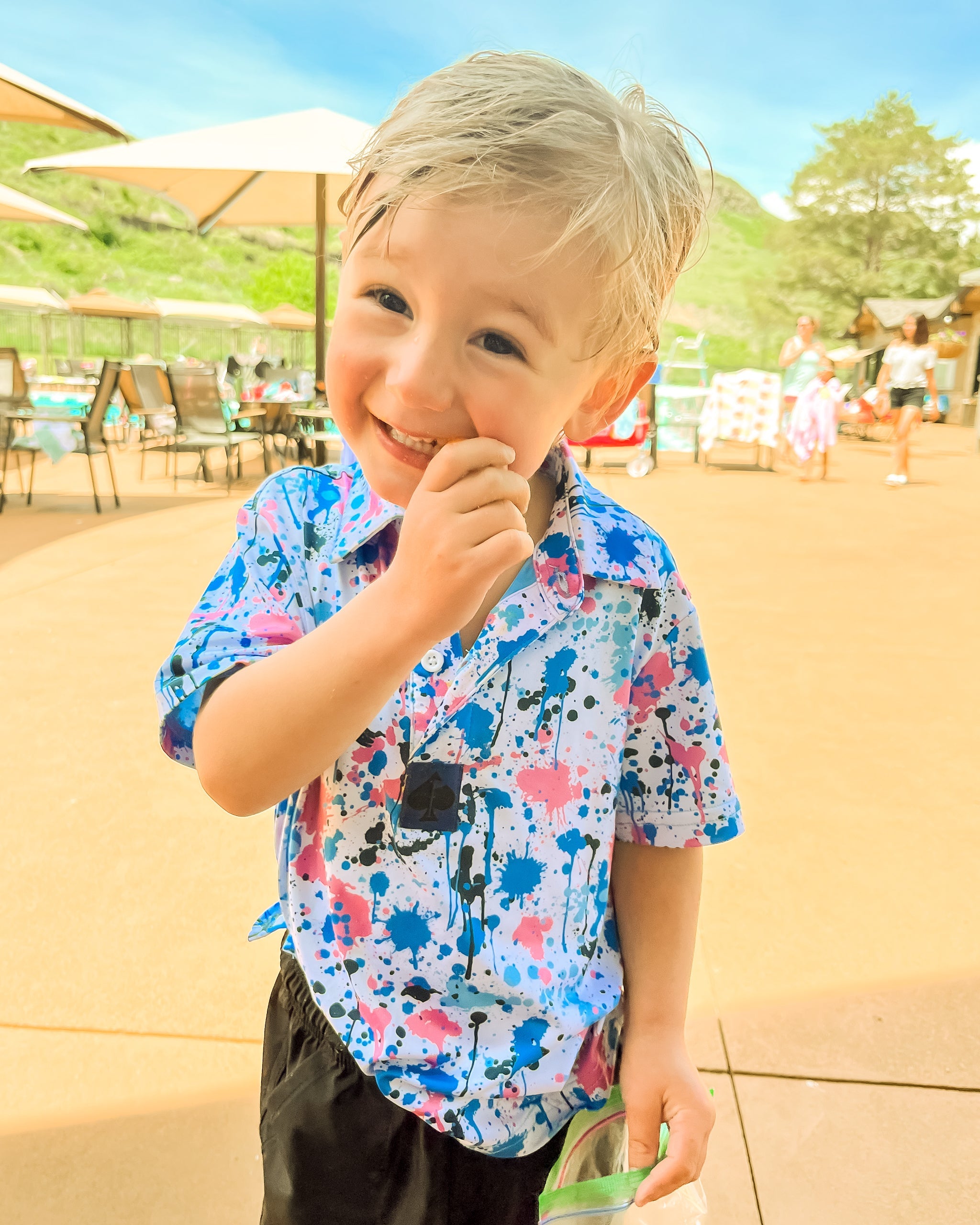 Youth in a colorful cotton candy drip polo shirt, smiling and enjoying a treat outdoors by a poolside.