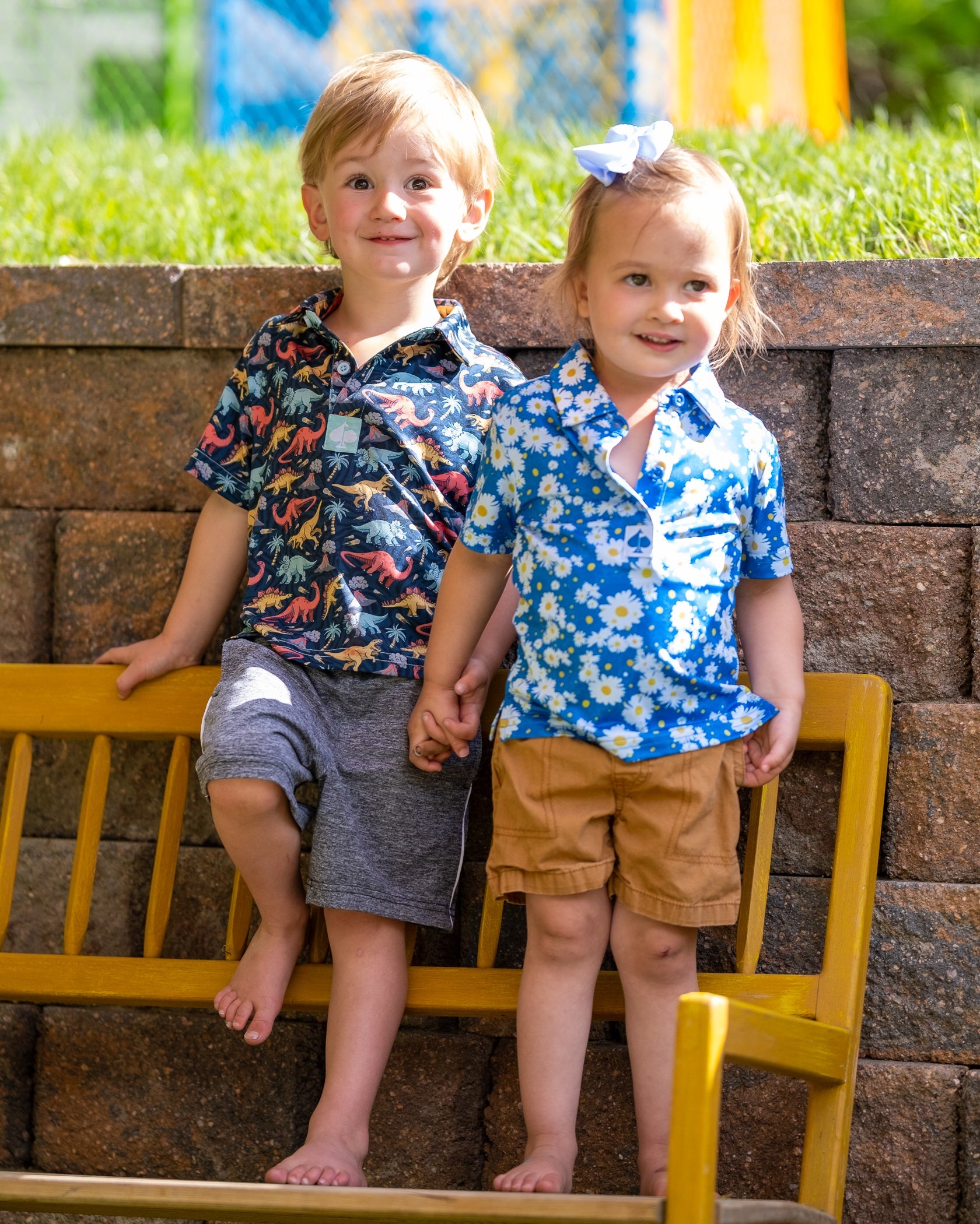 Two children stand on a bench, wearing colorful youth polos. One features a dinosaur print, and the other has a floral design.