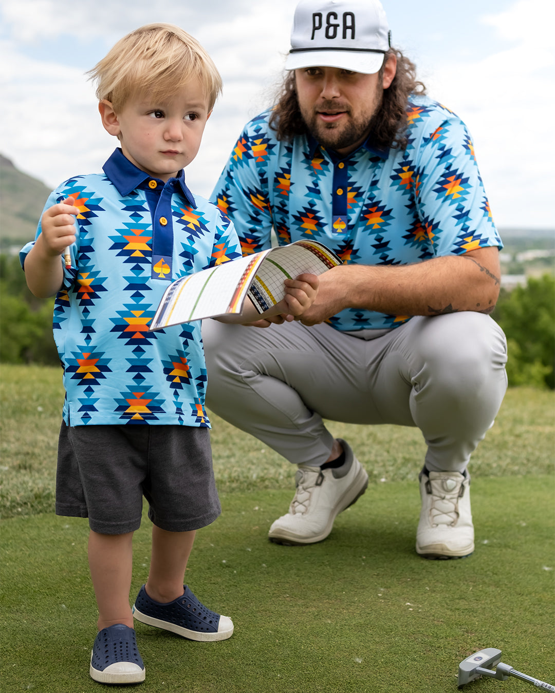 Youth in a colorful Southwest Sunset Polo stands on a golf course, while an adult reviews a scorecard nearby.