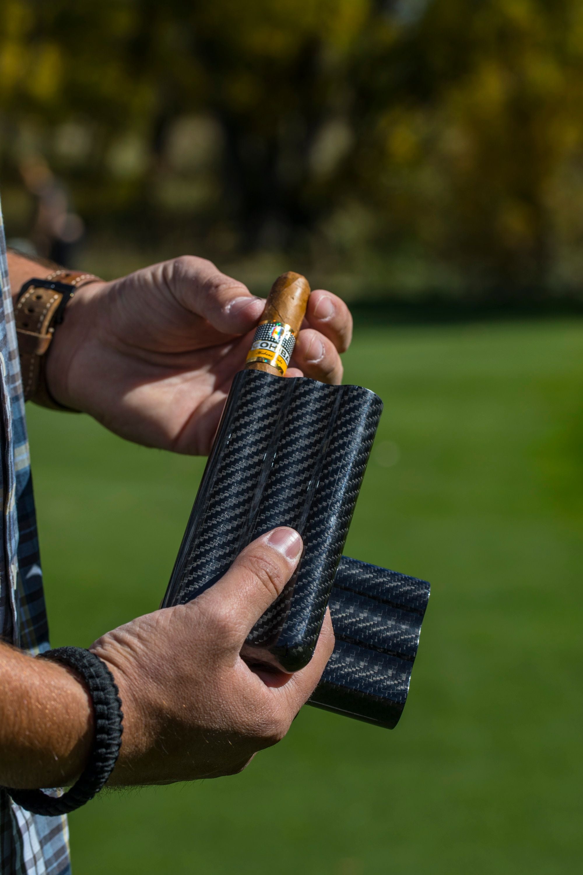 Carbon fiber cigar case with three slots, held by a person outdoors, showcasing a cigar being inserted.