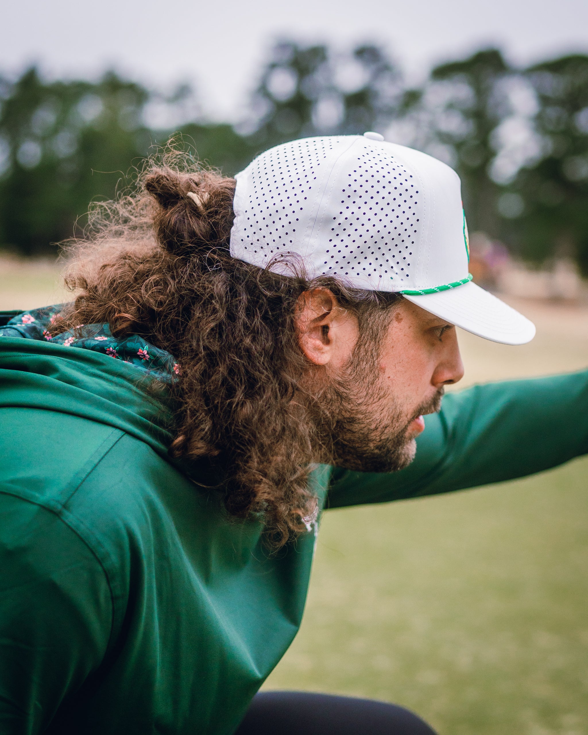 Performance Golf Hoodie in Dark Azalea worn by a man with curly hair, focusing intently on the golf course.