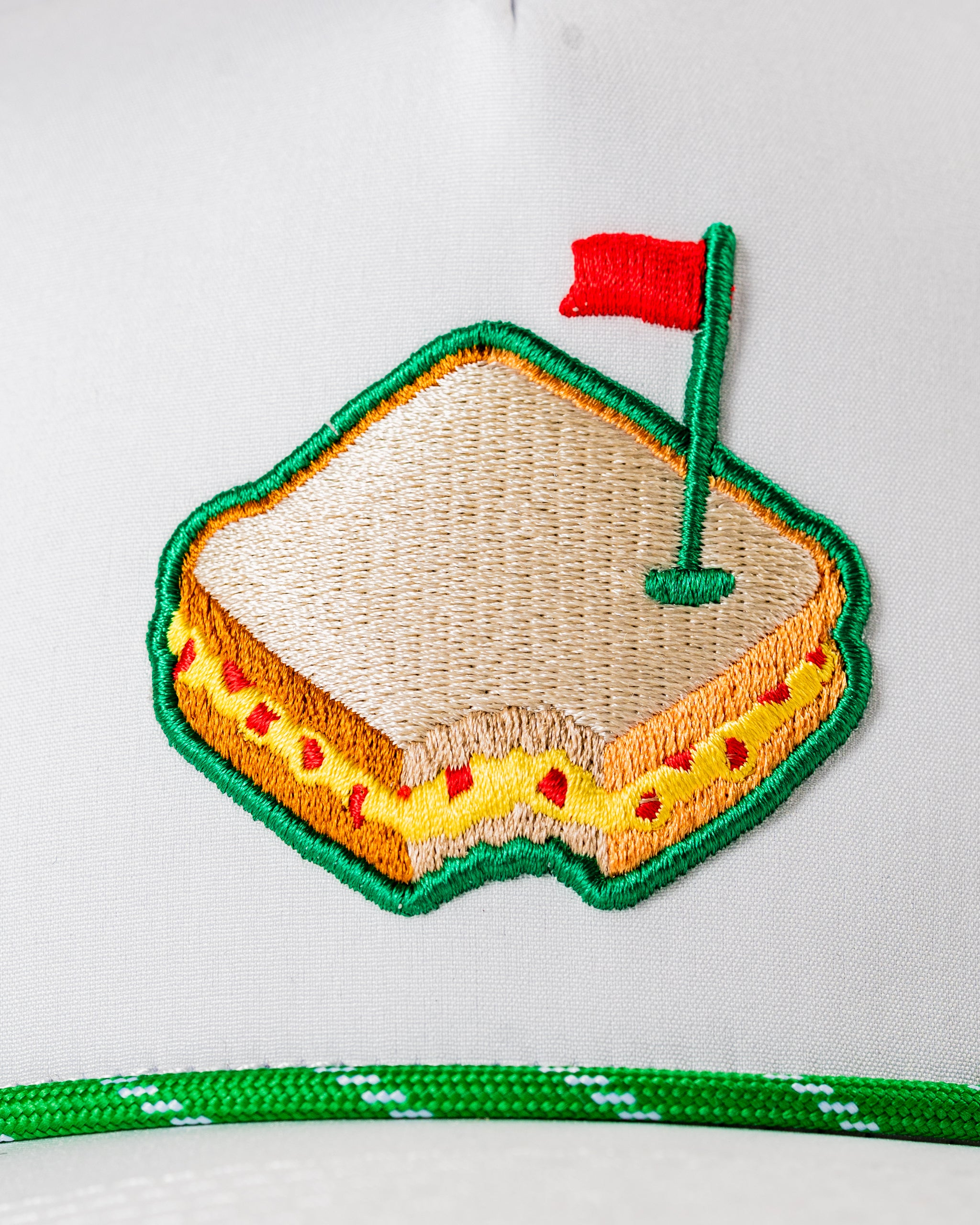 Perforated white rope hat featuring an embroidered pimento cheese sandwich with a golf flag. Ideal for stylish golfers.
