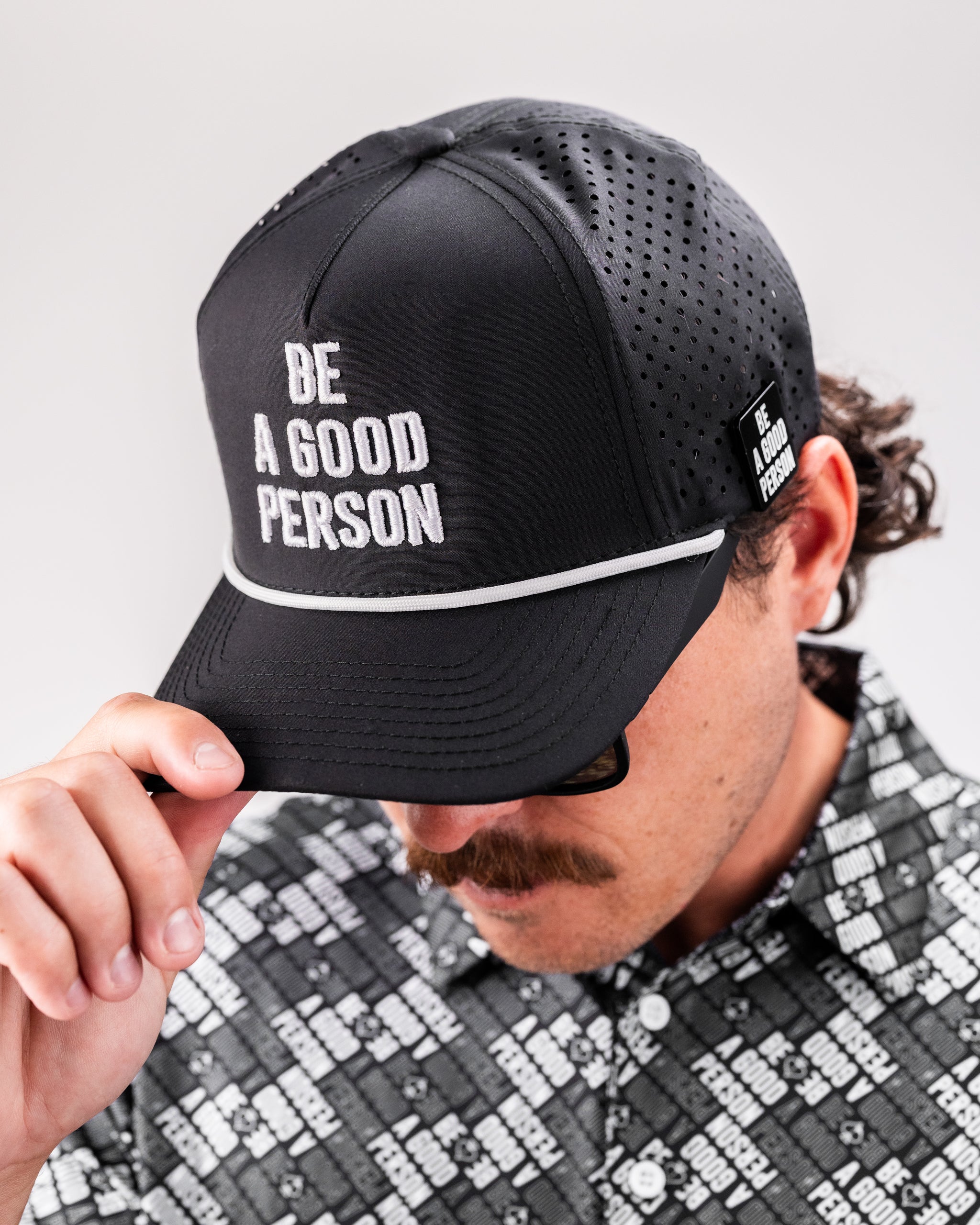 Black perforated rope hat featuring "Be A Good Person" embroidered on the front, worn by a man adjusting the brim.