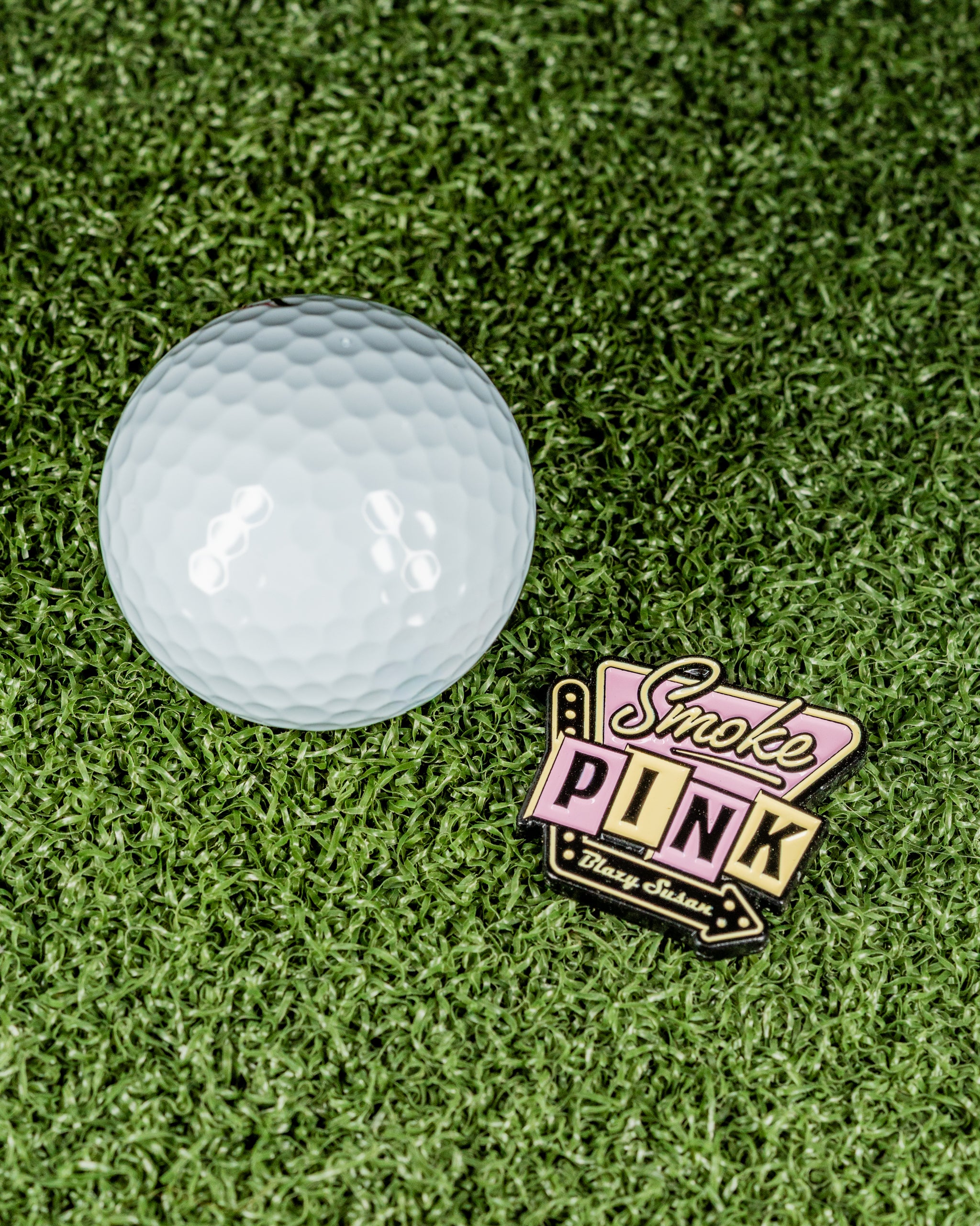 Blazy Susan Smoke Pink ball marker beside a white golf ball on green turf, showcasing a bold and vibrant design.