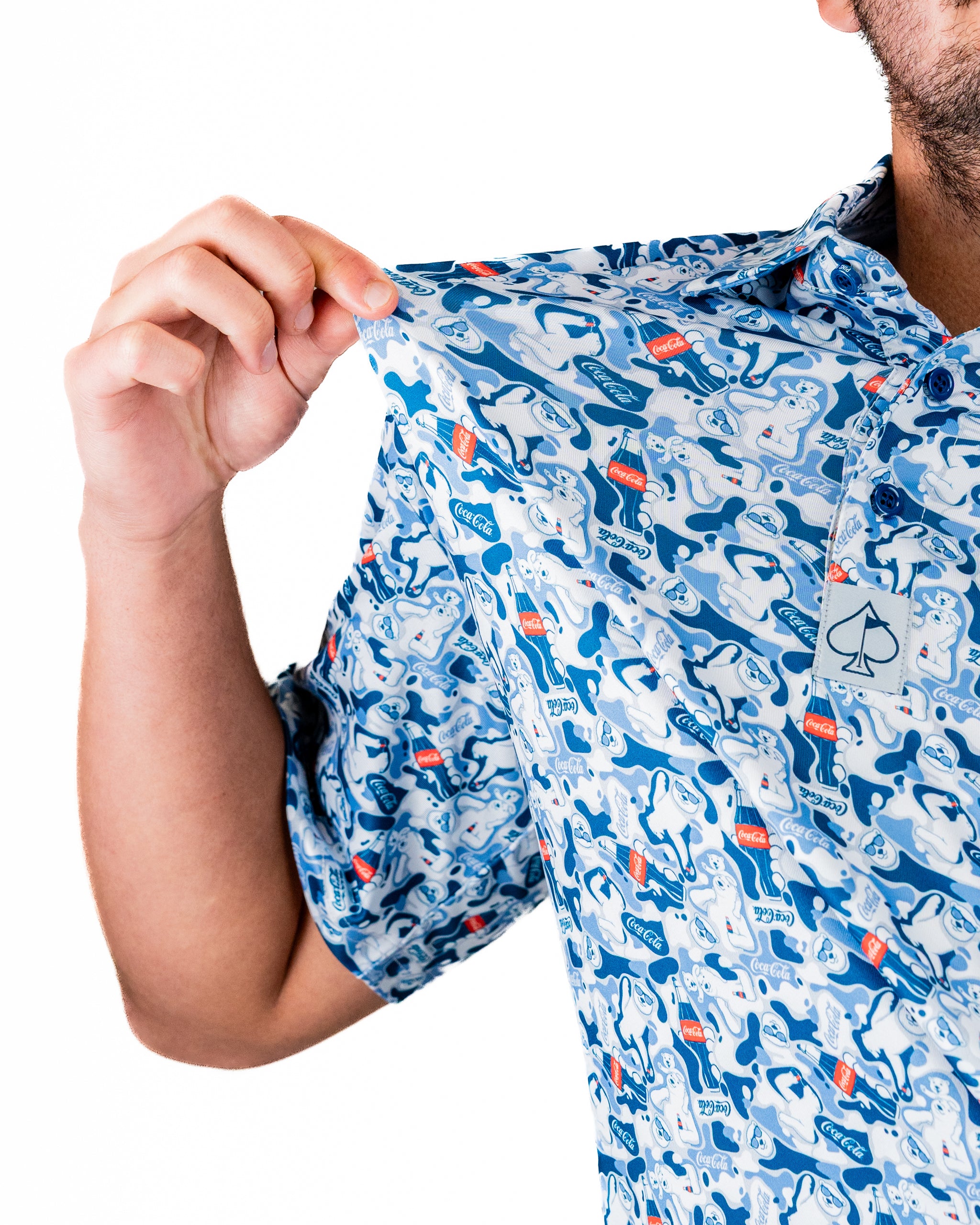 A person holds up a short-sleeve shirt featuring a playful Coca-Cola pattern with colorful graphics on a light blue background.