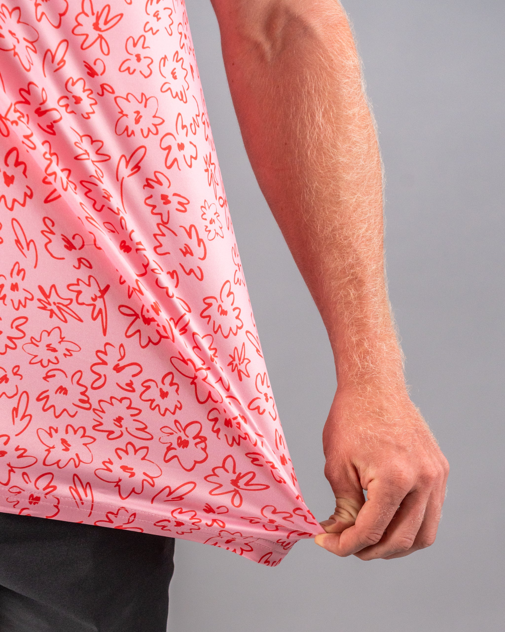 Close-up of a hand pulling the hem of a pink golf shirt featuring a playful flower doodle pattern in red.