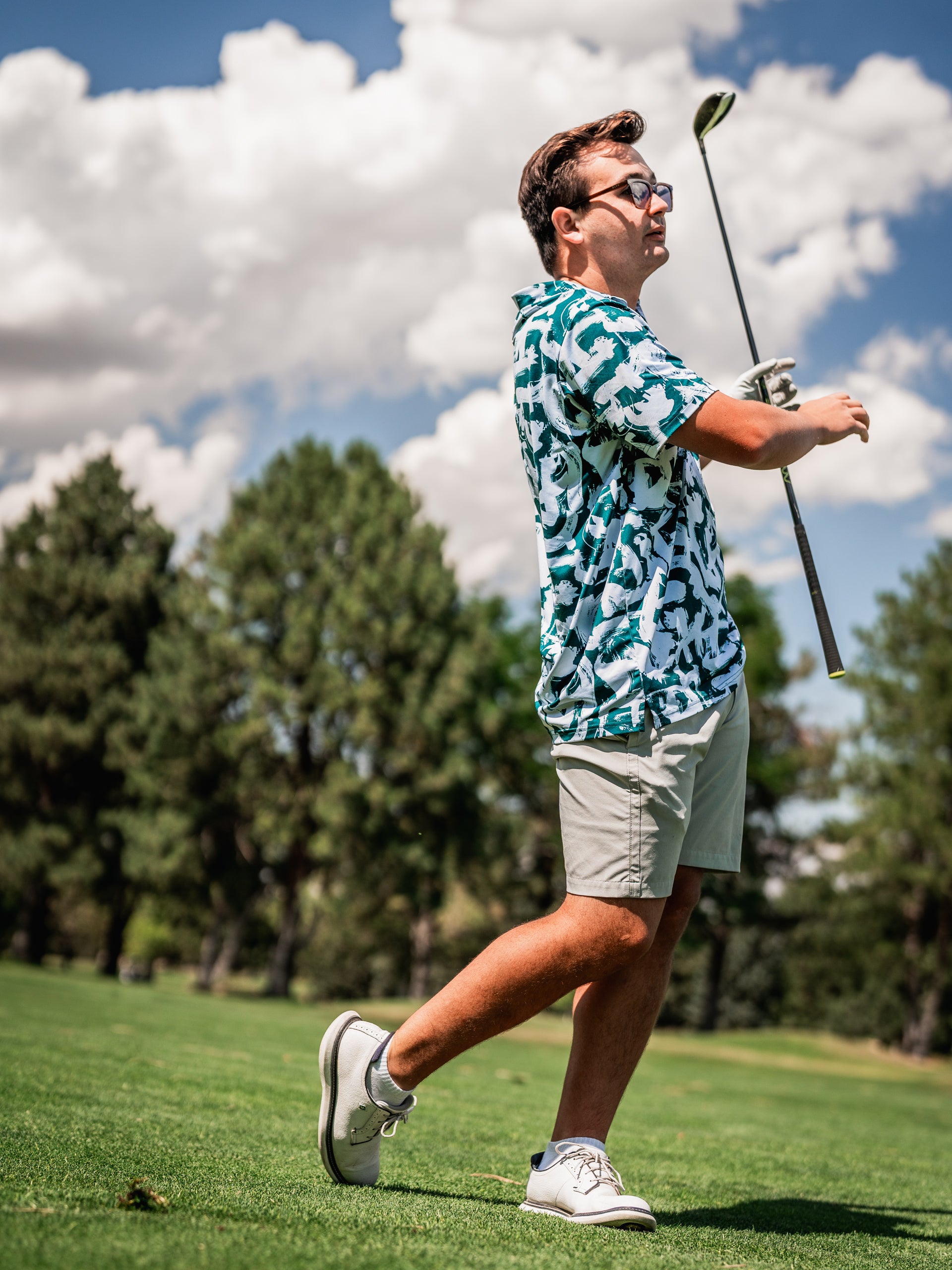 A golfer in a vibrant green paint strokes patterned shirt swings a club on a sunny course, showcasing bold style.