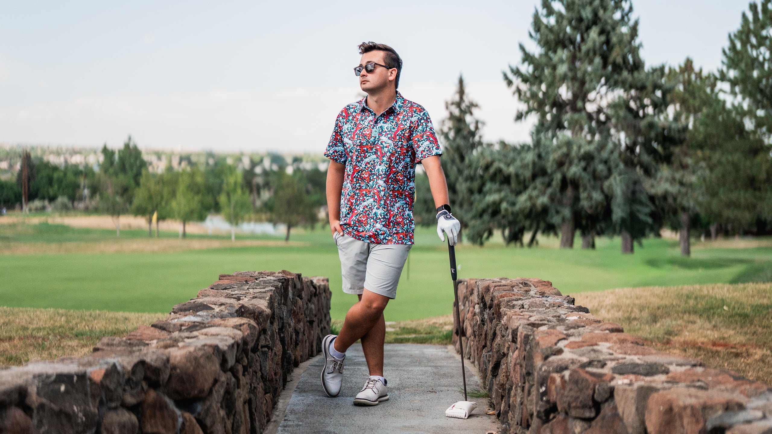 A golfer in a vibrant floral shirt and shorts stands confidently on a stone path, holding a golf club, with a scenic course in the background.