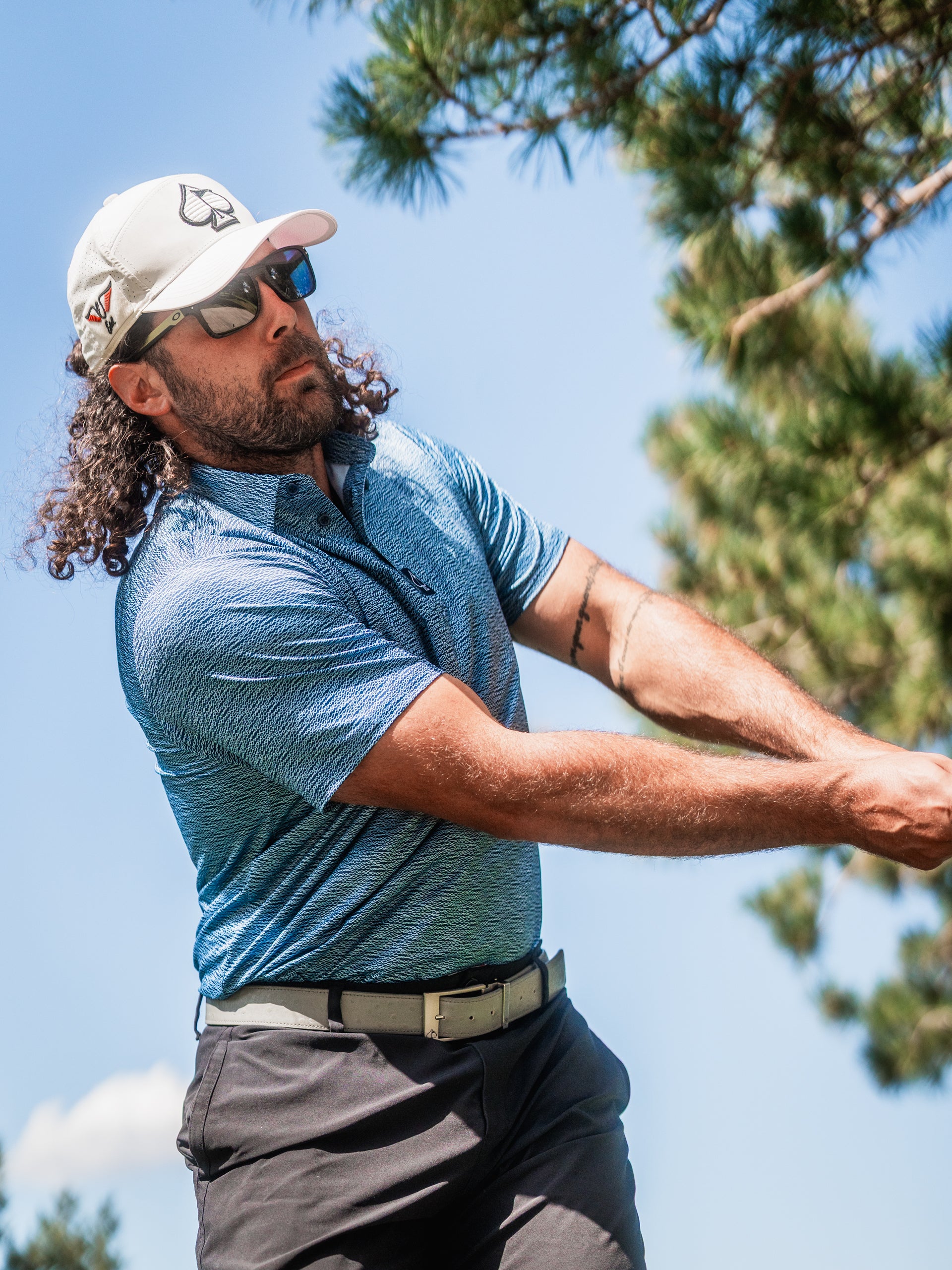 A golfer swings a club while wearing a blue patterned polo shirt, sunglasses, and a cap, set against a sunny backdrop.