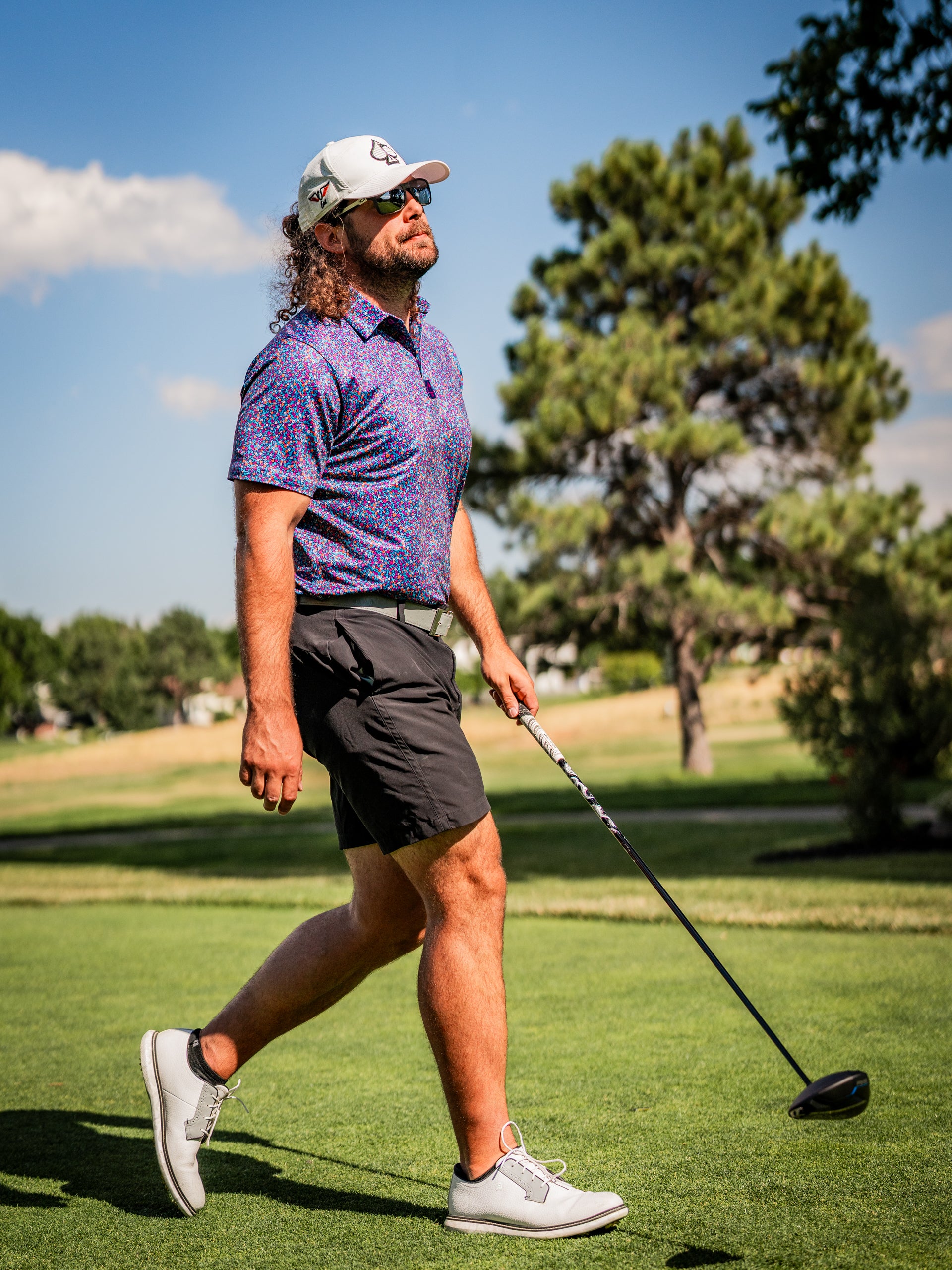 A golfer in a lavender floral polo shirt and black shorts walks confidently on the course, holding a driver.