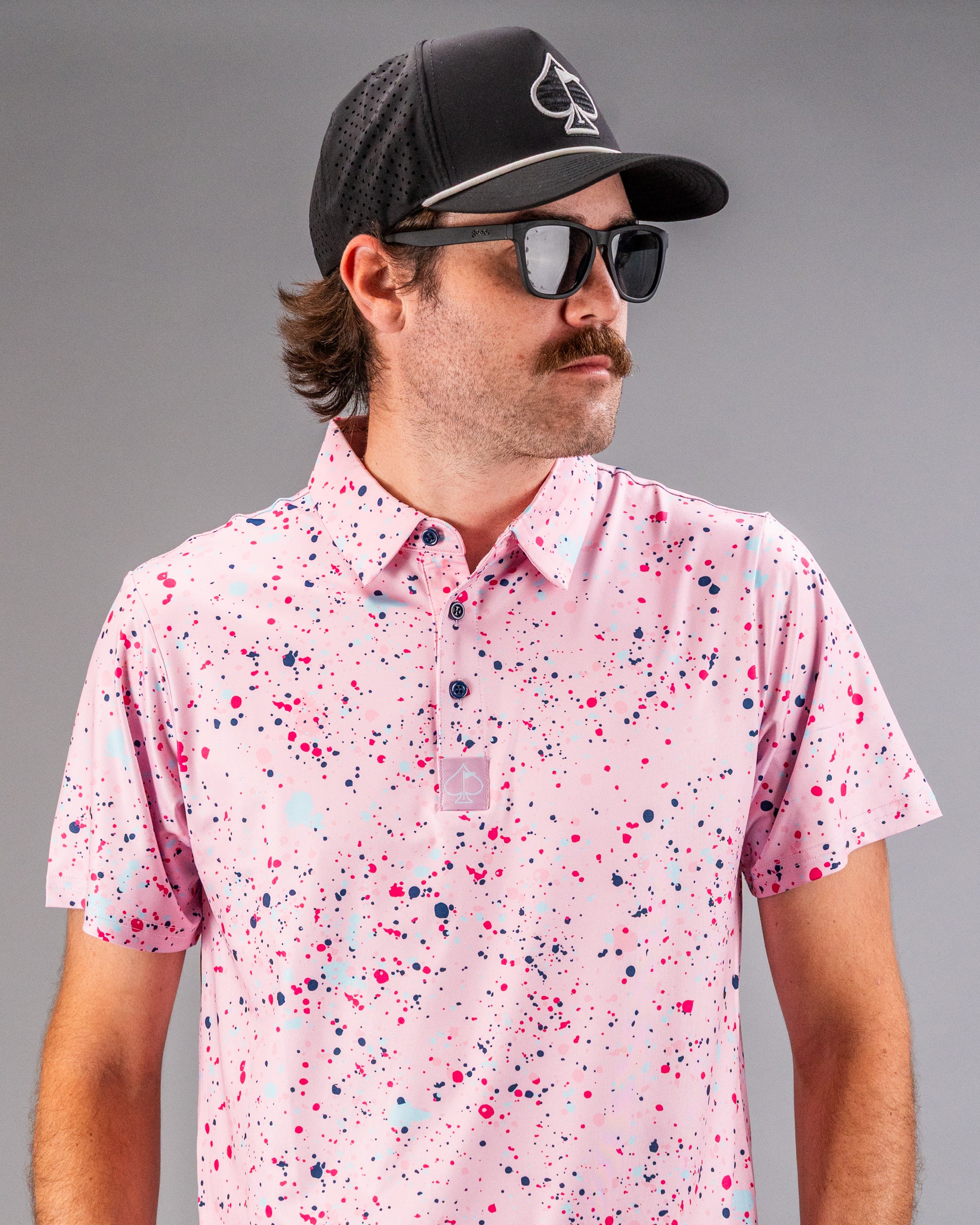 Stylish golfer wearing a pink splatter polo shirt with a black cap and sunglasses, showcasing bold golf fashion.