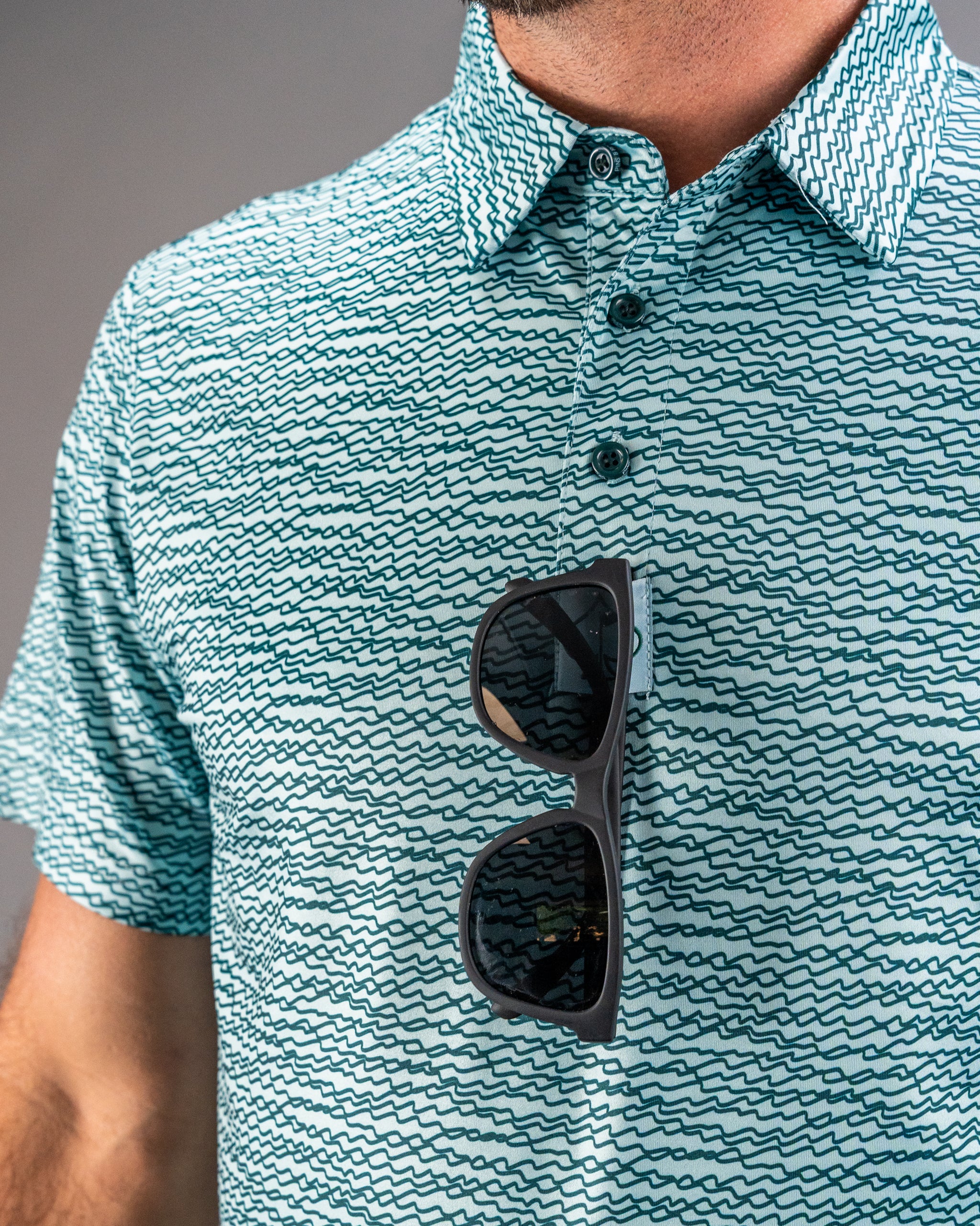 Teal golf polo with a squiggle pattern, featuring a collar and sunglasses resting on the chest.