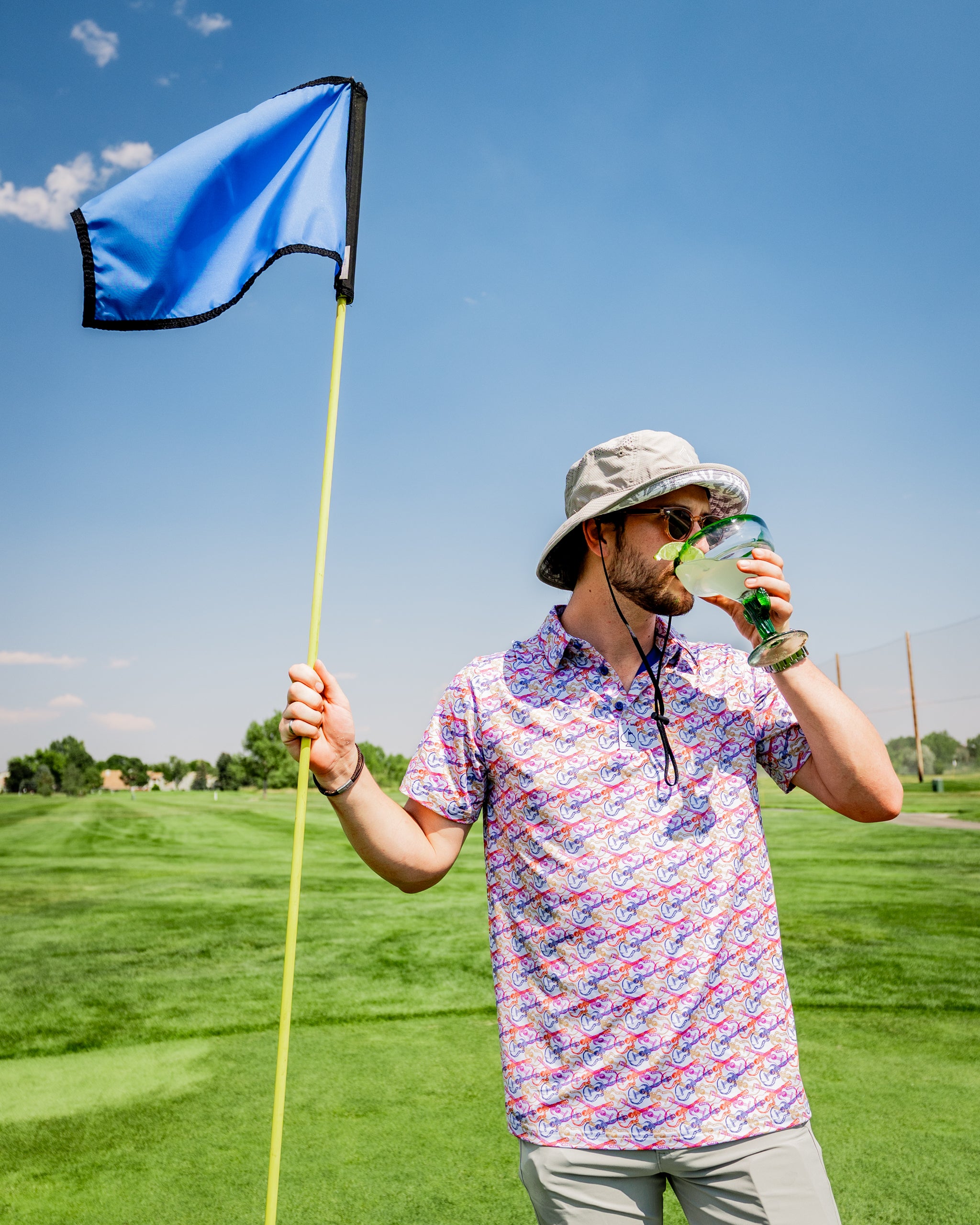 Man on a golf course holds a blue flag and drinks from a large glass, wearing a colorful Margaritaville shirt.